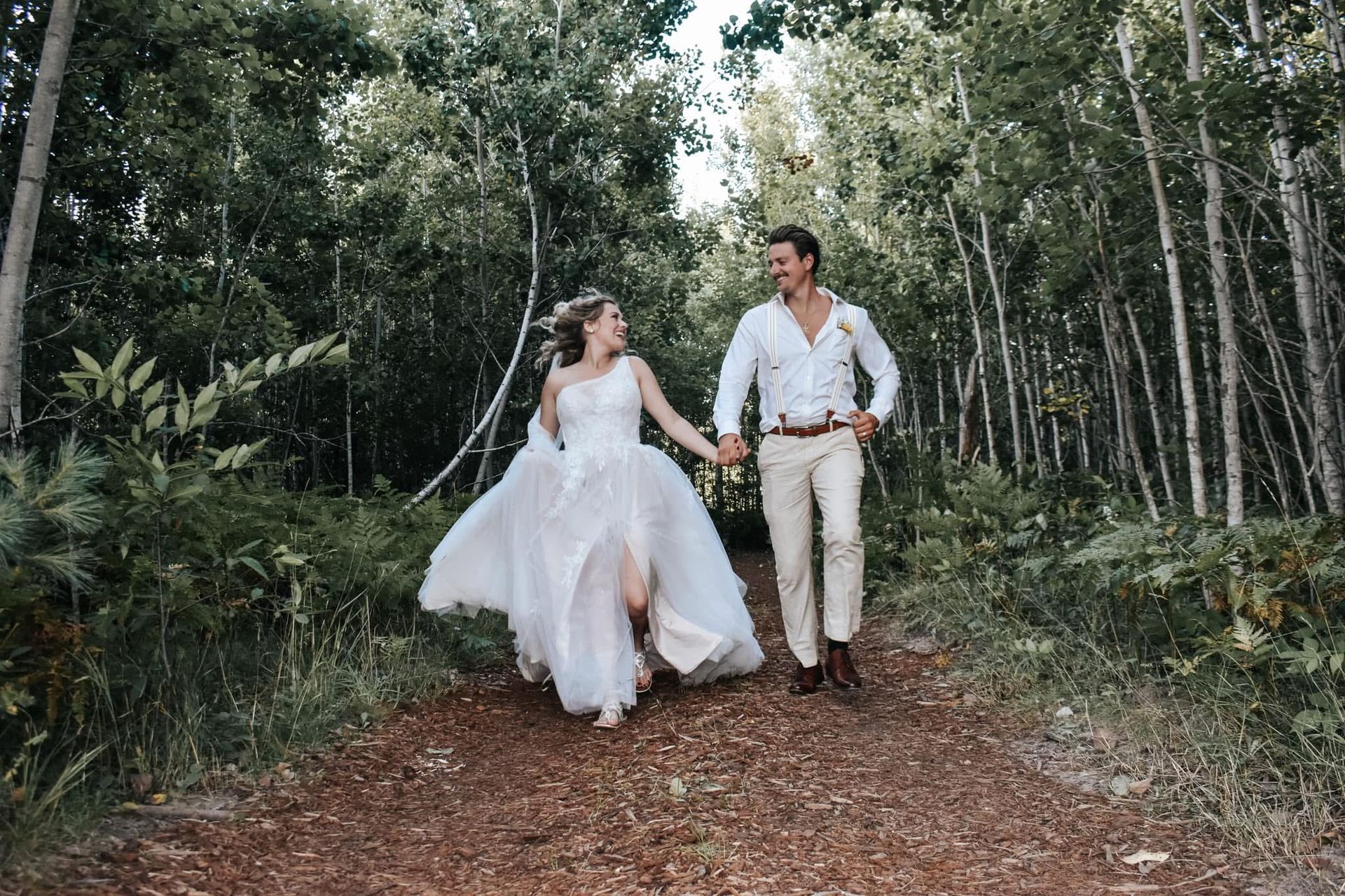 A bride and groom are walking down a path in the woods holding hands.
