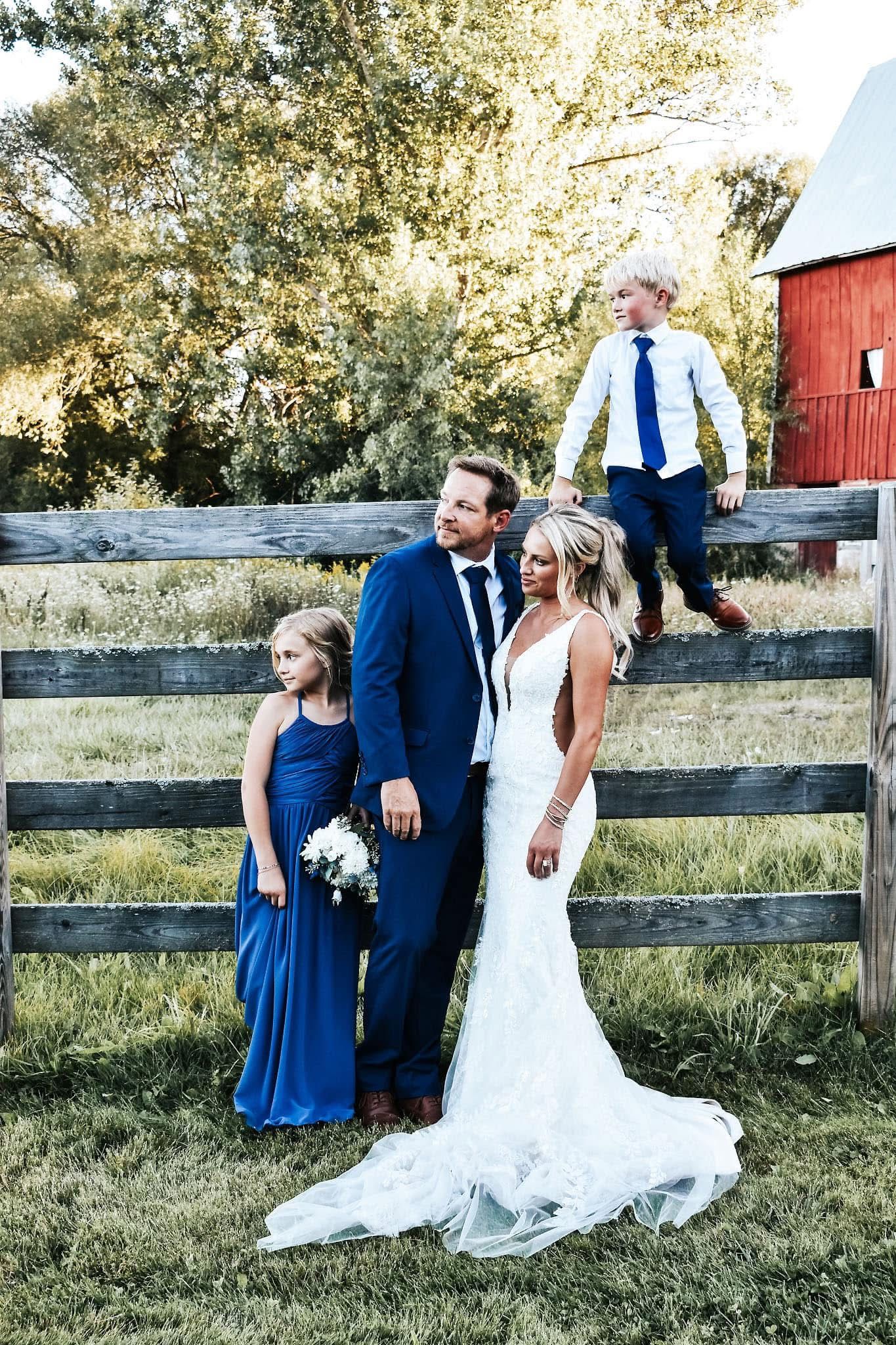 A bride and groom are posing for a picture with their children in front of a barn.
