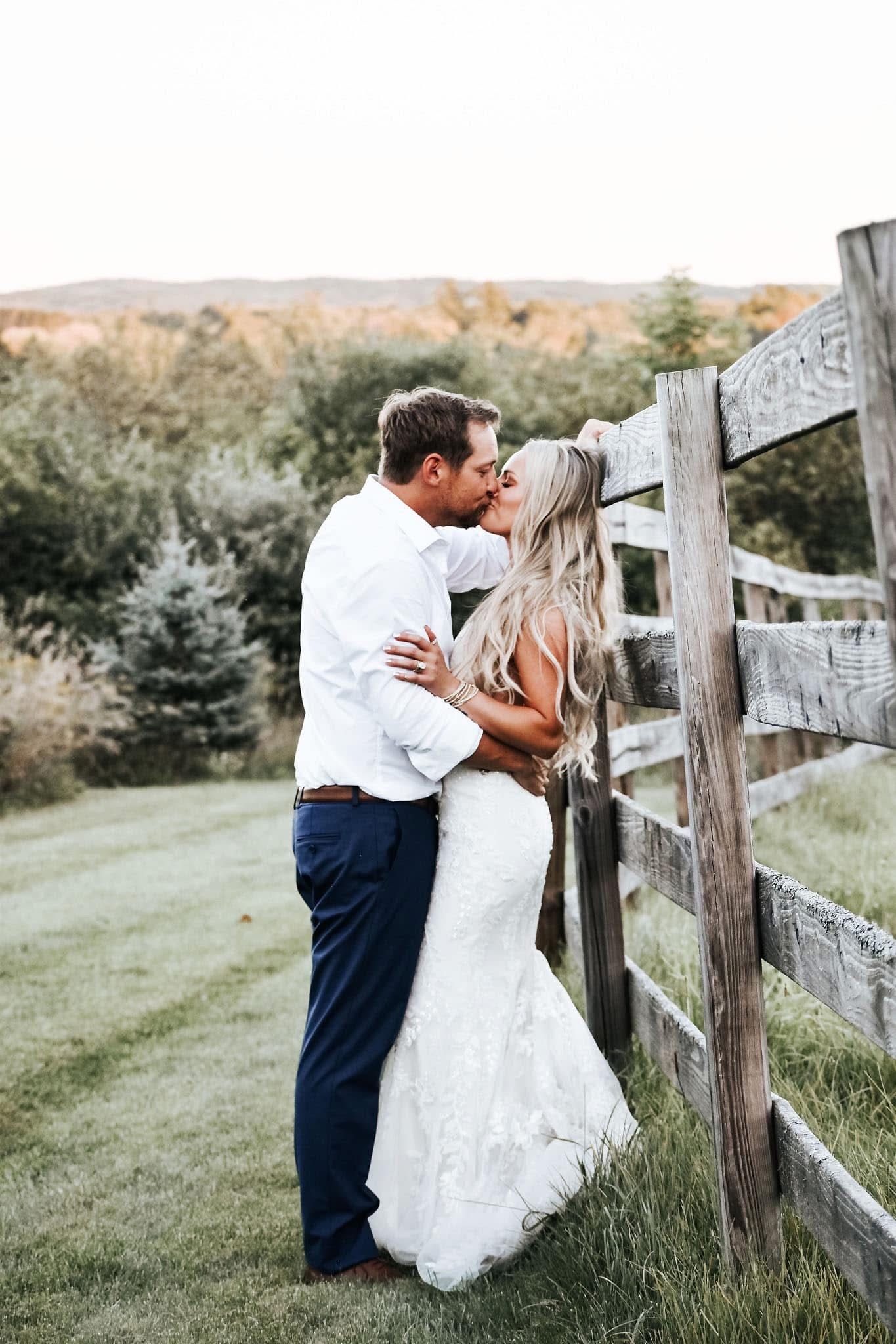A bride and groom are kissing next to a wooden fence.