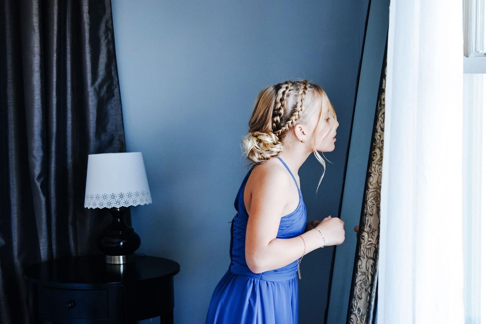 A little girl in a blue dress is looking at herself in the mirror.