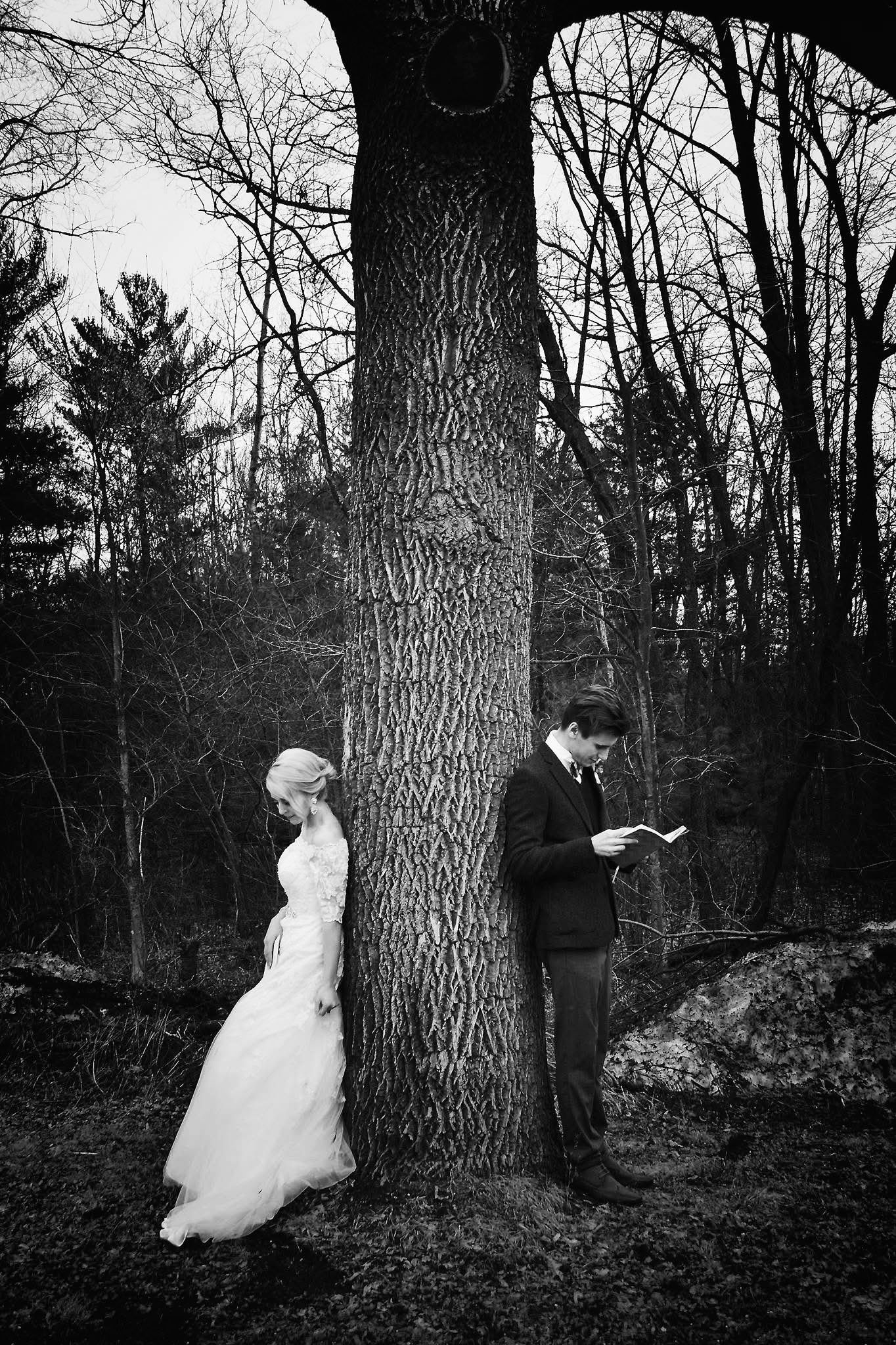 A black and white photo of a bride and groom standing next to a tree.