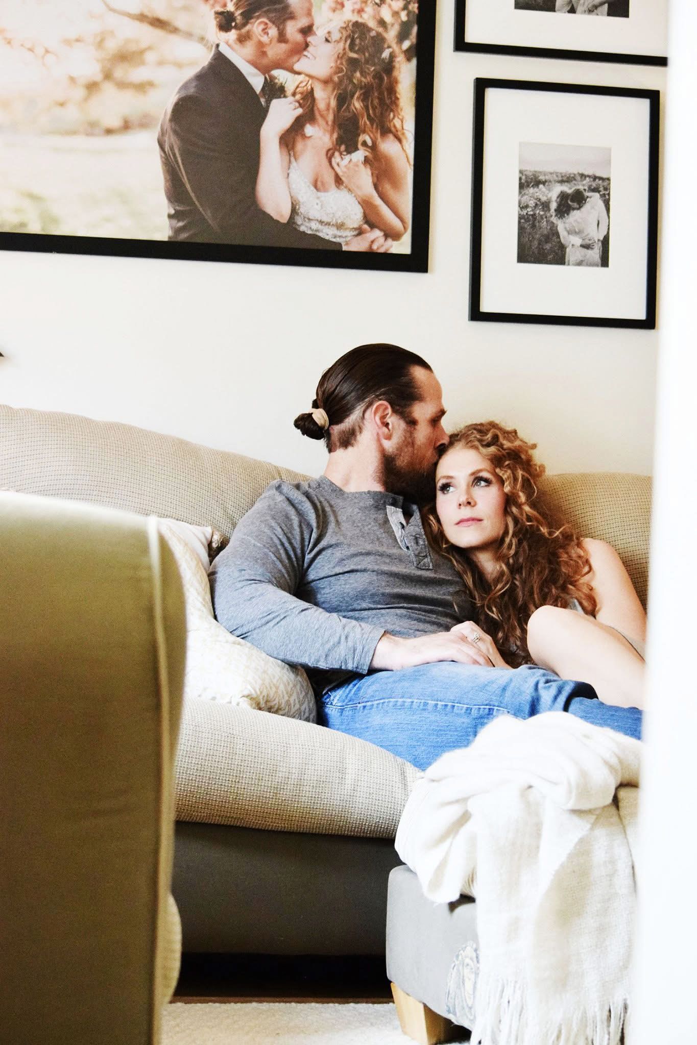 A man and woman are sitting on a couch with a picture of a bride and groom on the wall behind them.