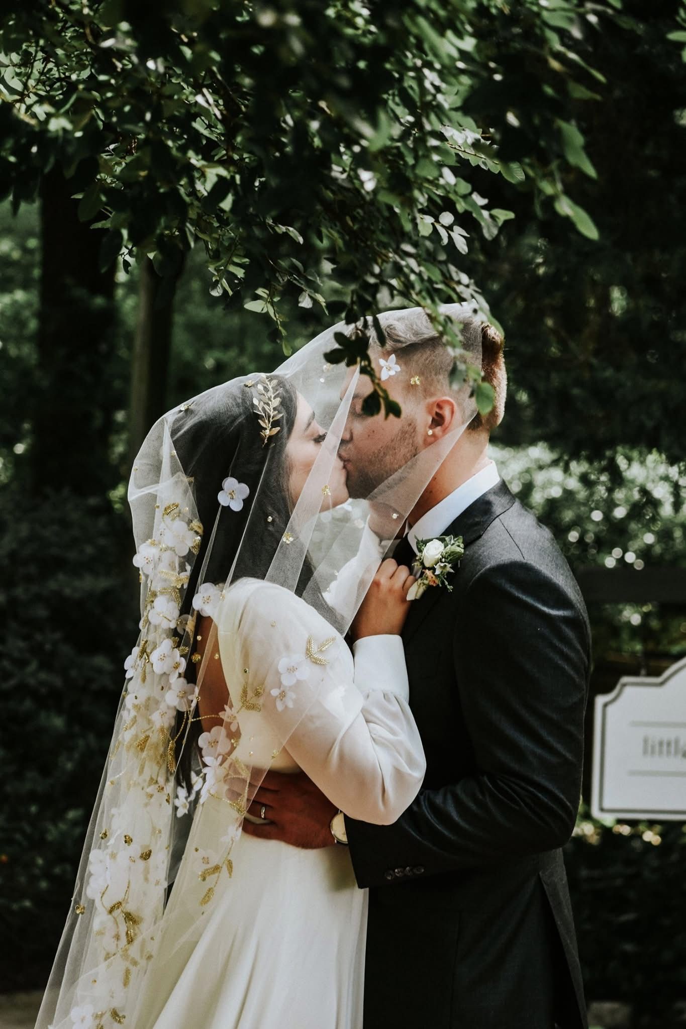 A bride and groom kissing under a veil in a park.