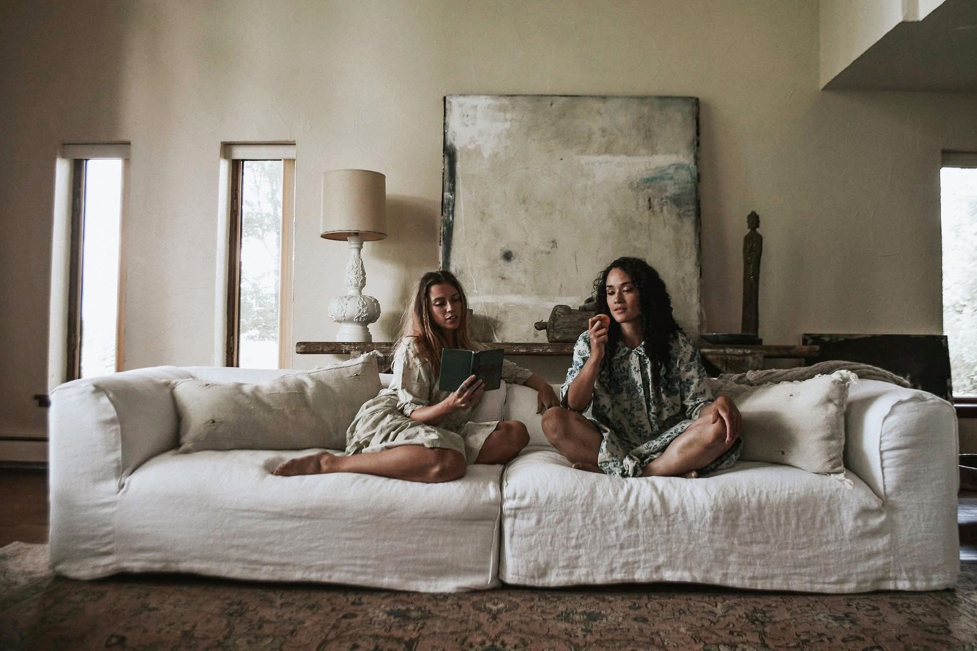 Two women are sitting on a white couch in a living room.