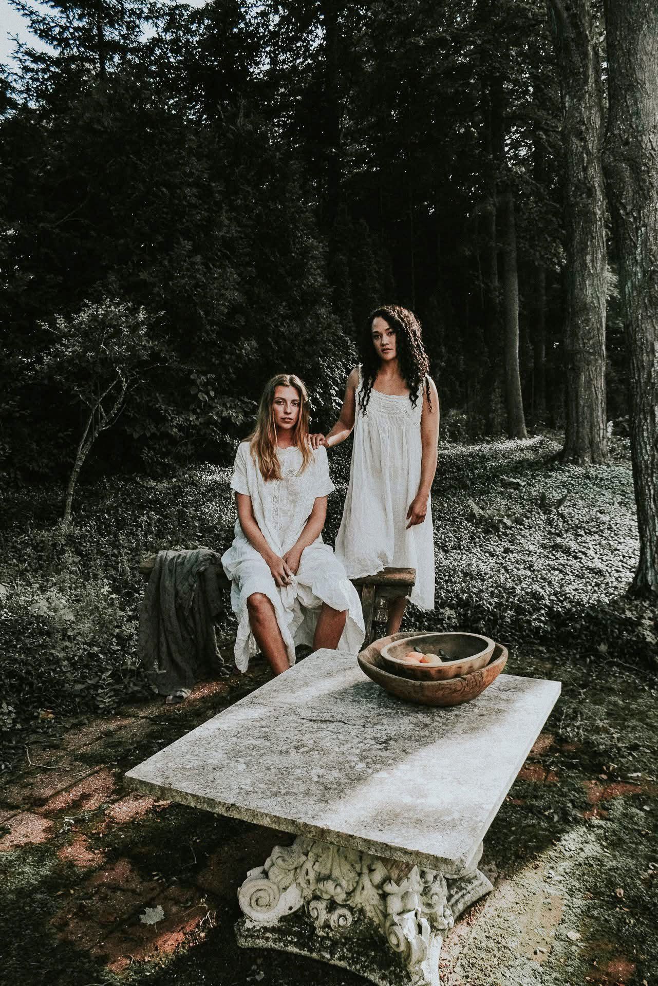 Two women in white dresses are sitting at a table in the woods.