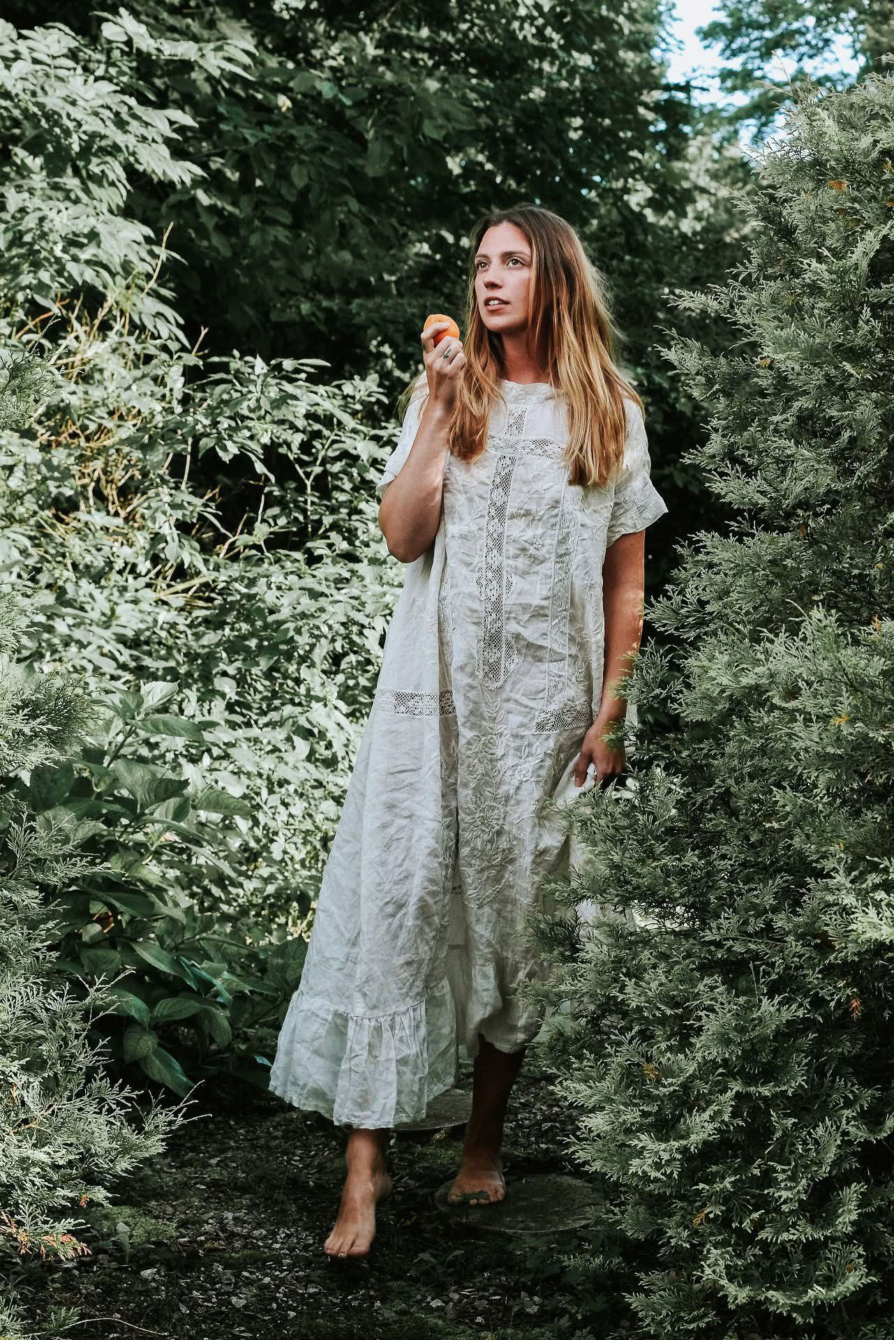A woman in a white lace dress is standing in the woods holding an apple.