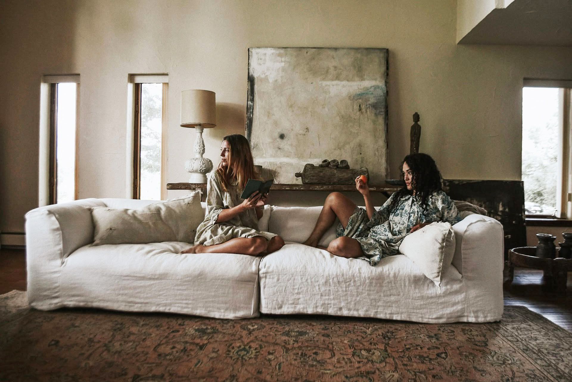 Two women are sitting on a white couch in a living room.