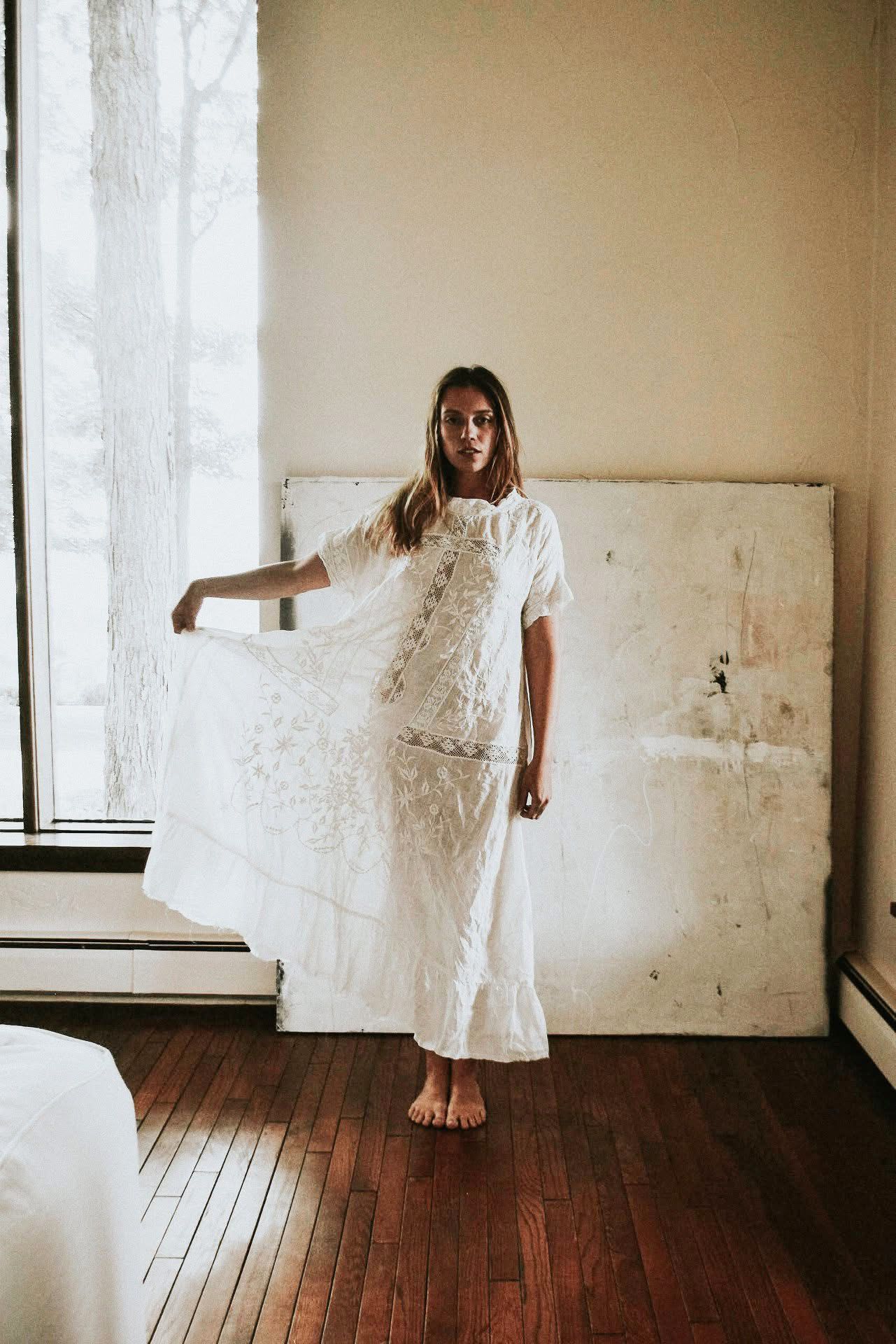 A woman in a white dress is standing in front of a window in a room.