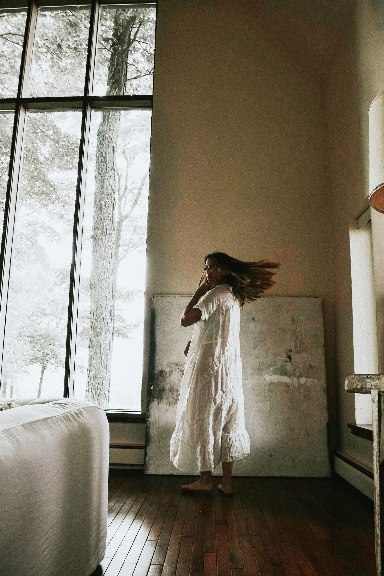 A woman in a white dress is standing in front of a large window in a living room.