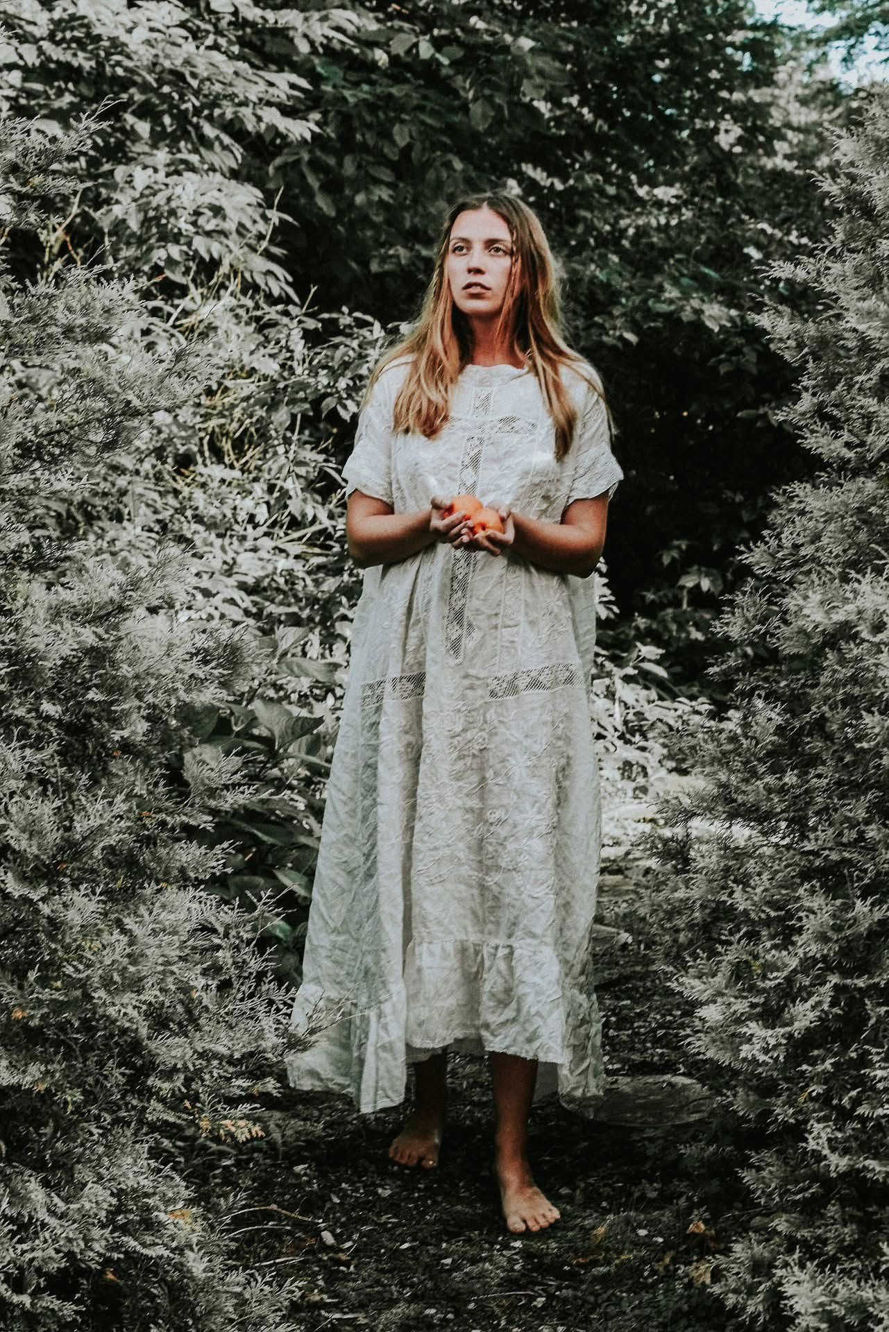A woman in a white dress is standing in the woods holding a flower.
