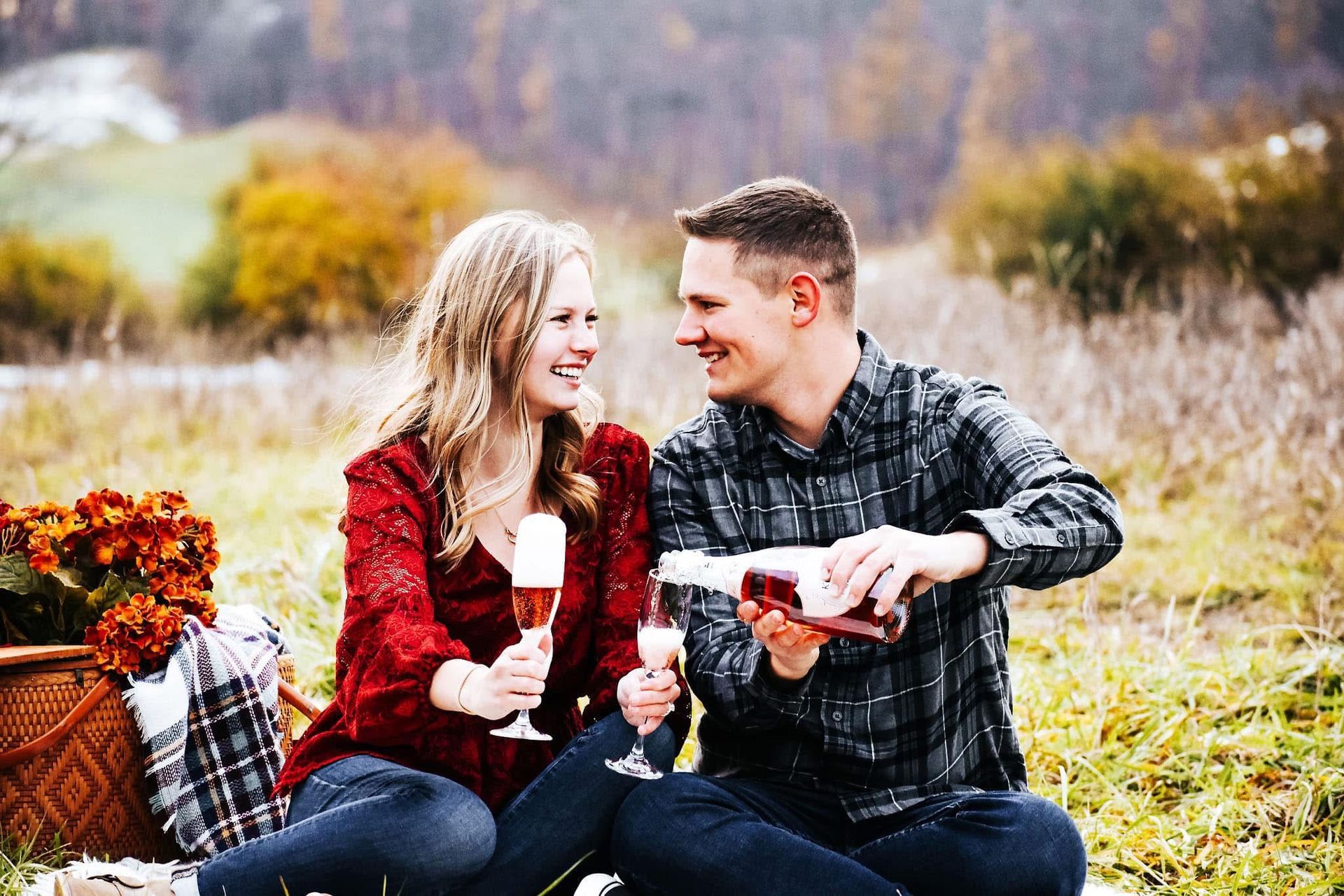 A man and a woman are sitting on a blanket in a field drinking wine.