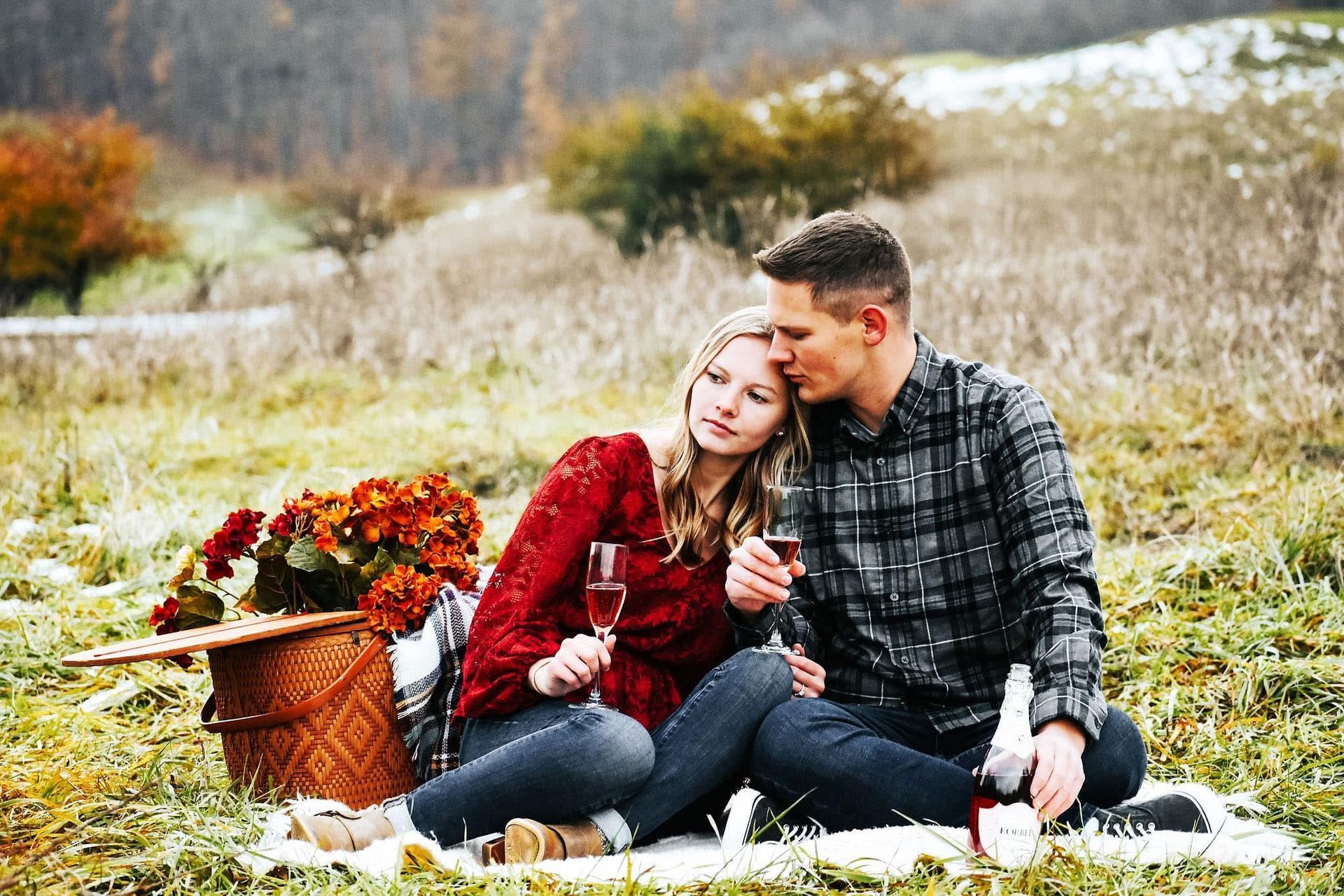A man and a woman are sitting on a blanket in a field drinking wine.
