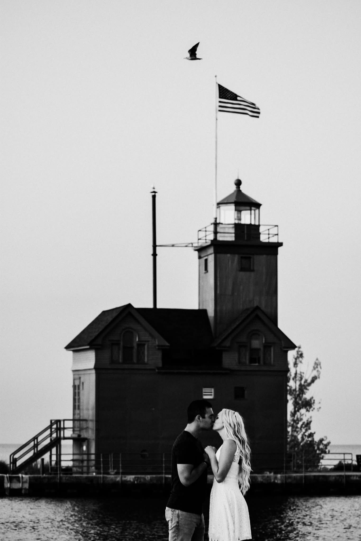 A black and white photo of a man and woman kissing in front of a lighthouse