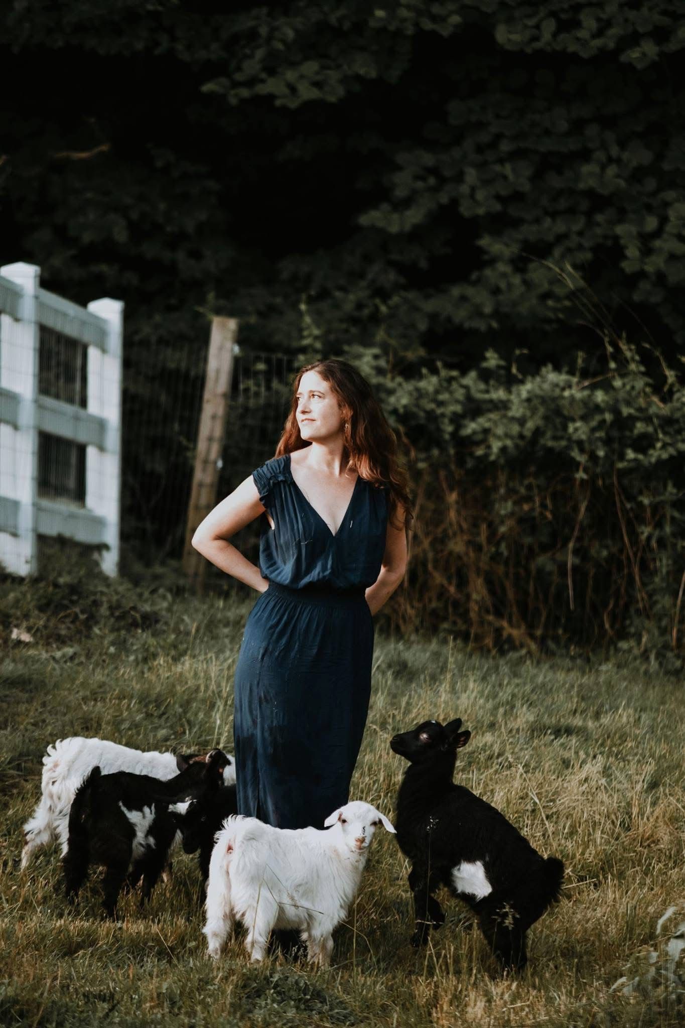 A woman in a blue dress is standing next to a herd of goats in a field.