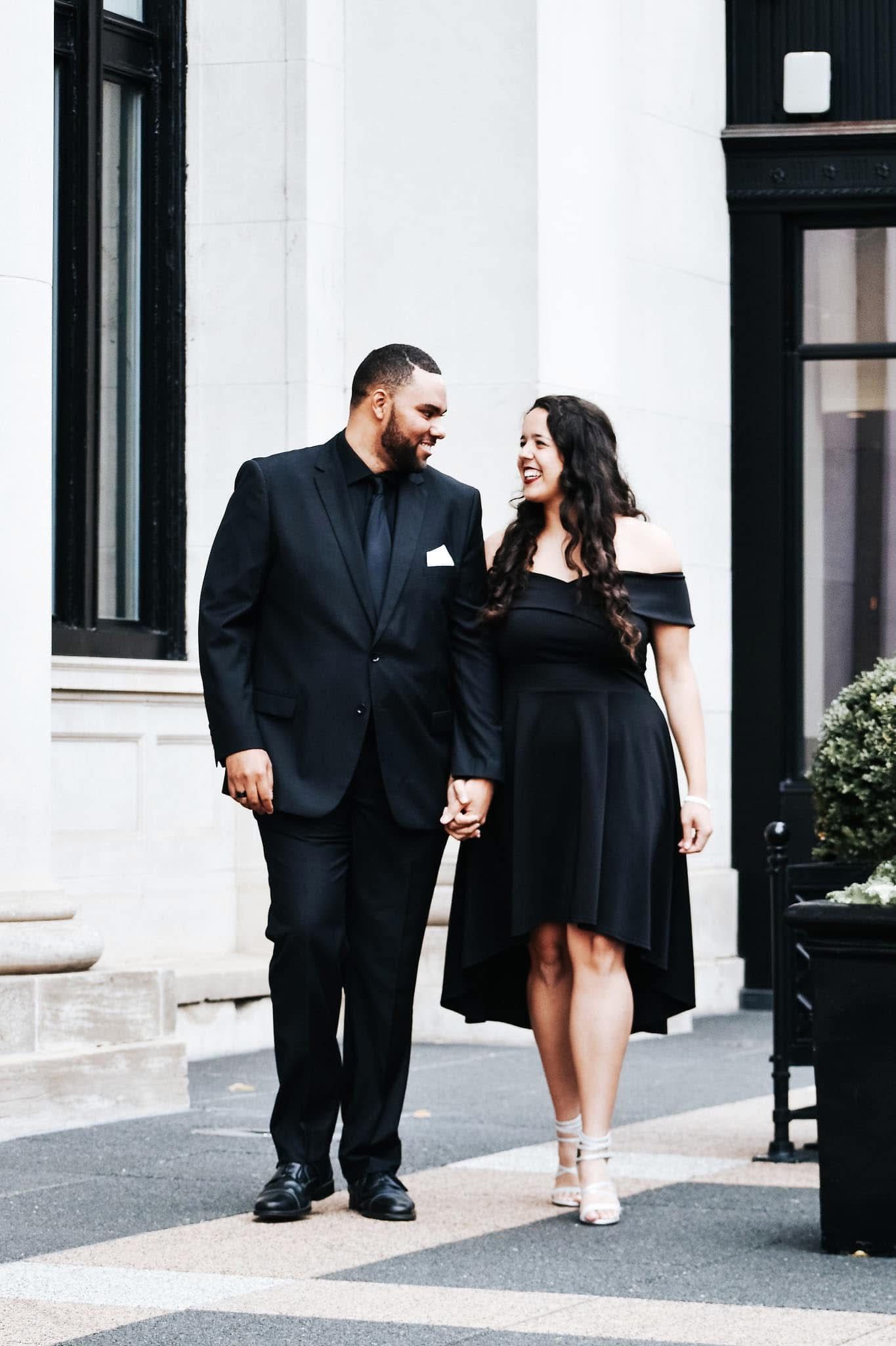 A man in a suit and a woman in a black dress are walking down a sidewalk holding hands.