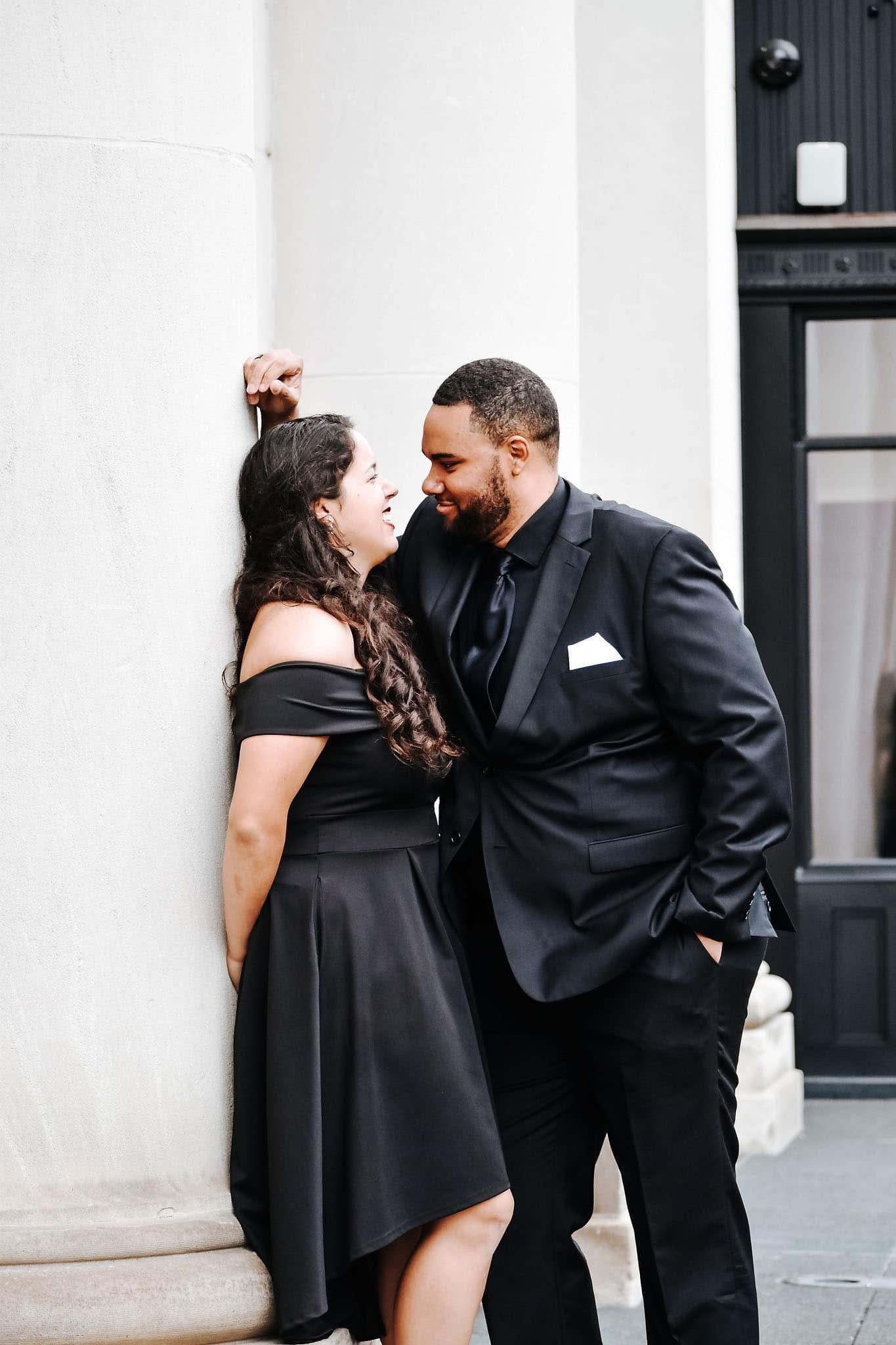 A man in a suit and a woman in a black dress are leaning against a wall.