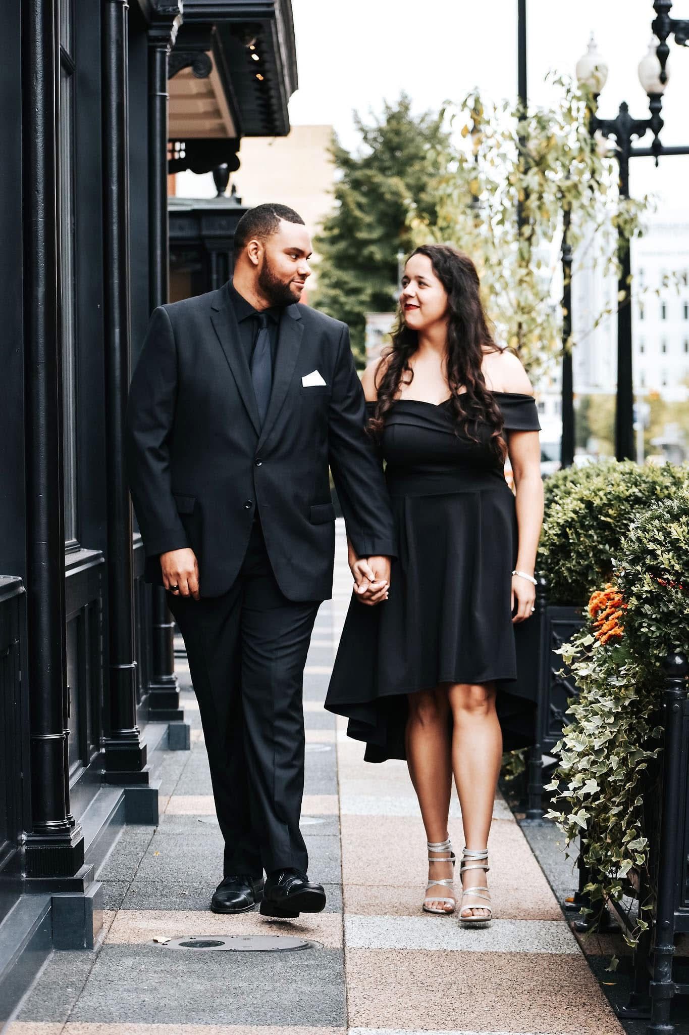 A man in a suit and a woman in a black dress are walking down a sidewalk holding hands.