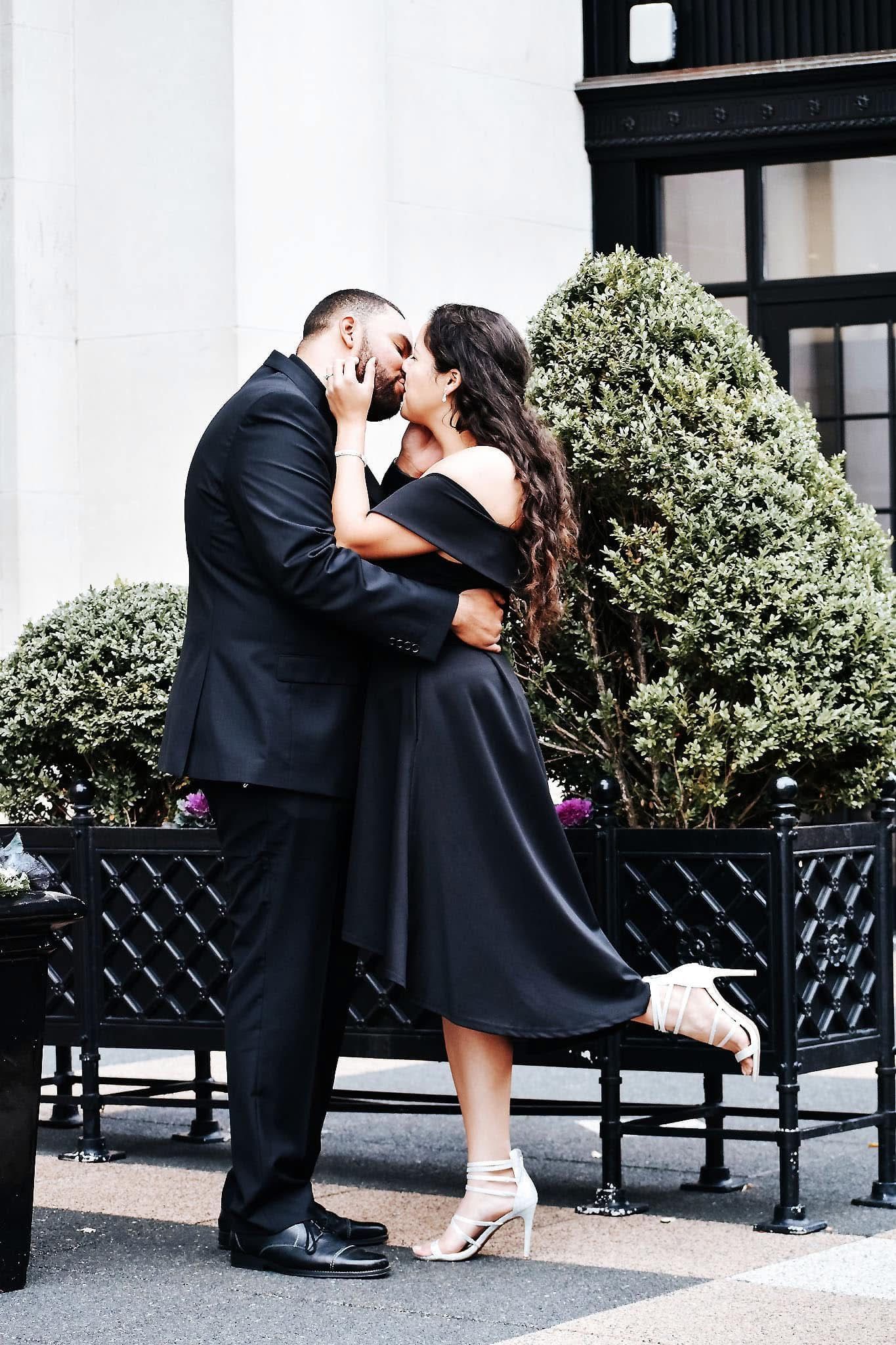 A man and a woman are kissing in front of a building . the woman is wearing a black dress.