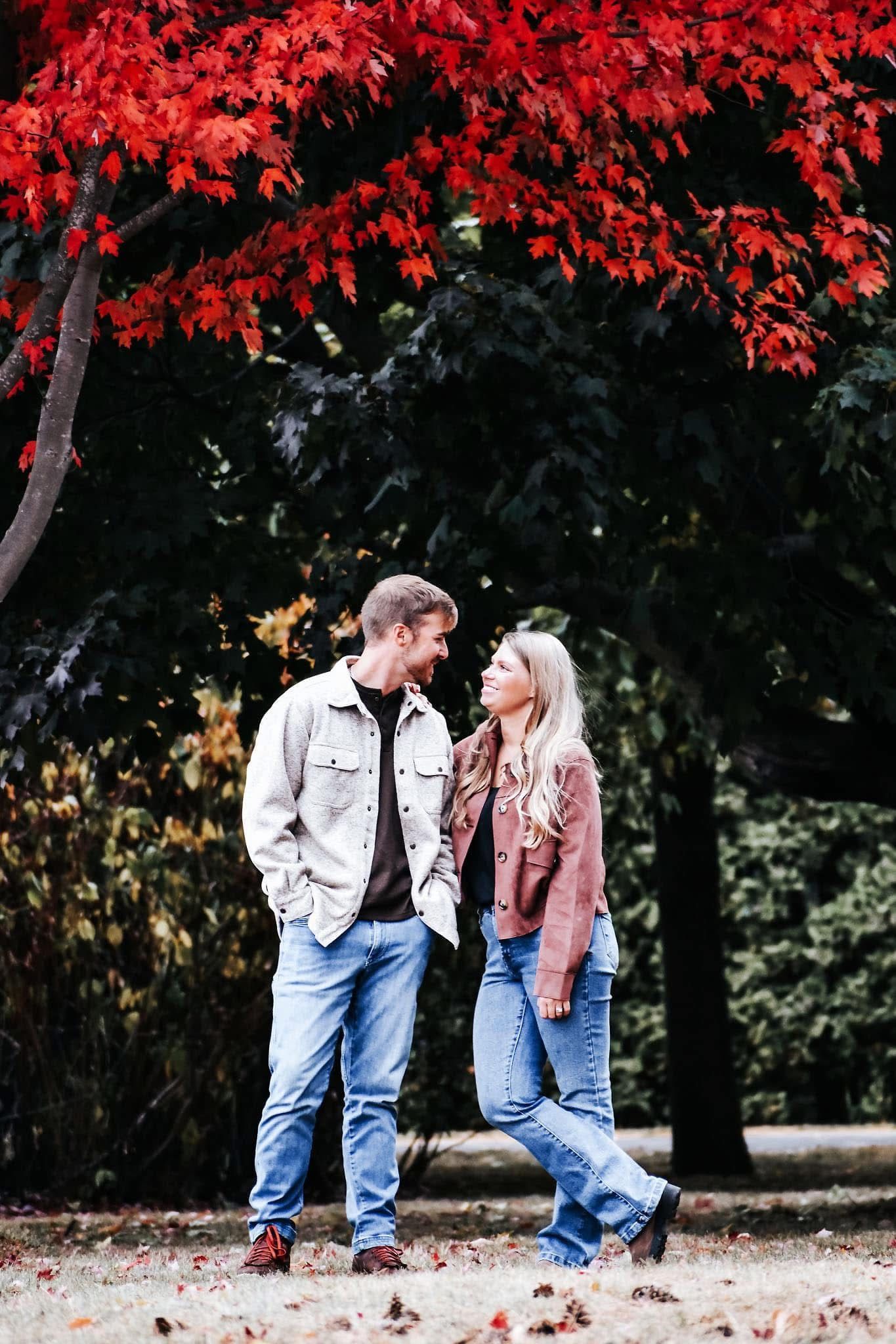 A man and a woman are standing next to each other in front of a tree with red leaves.