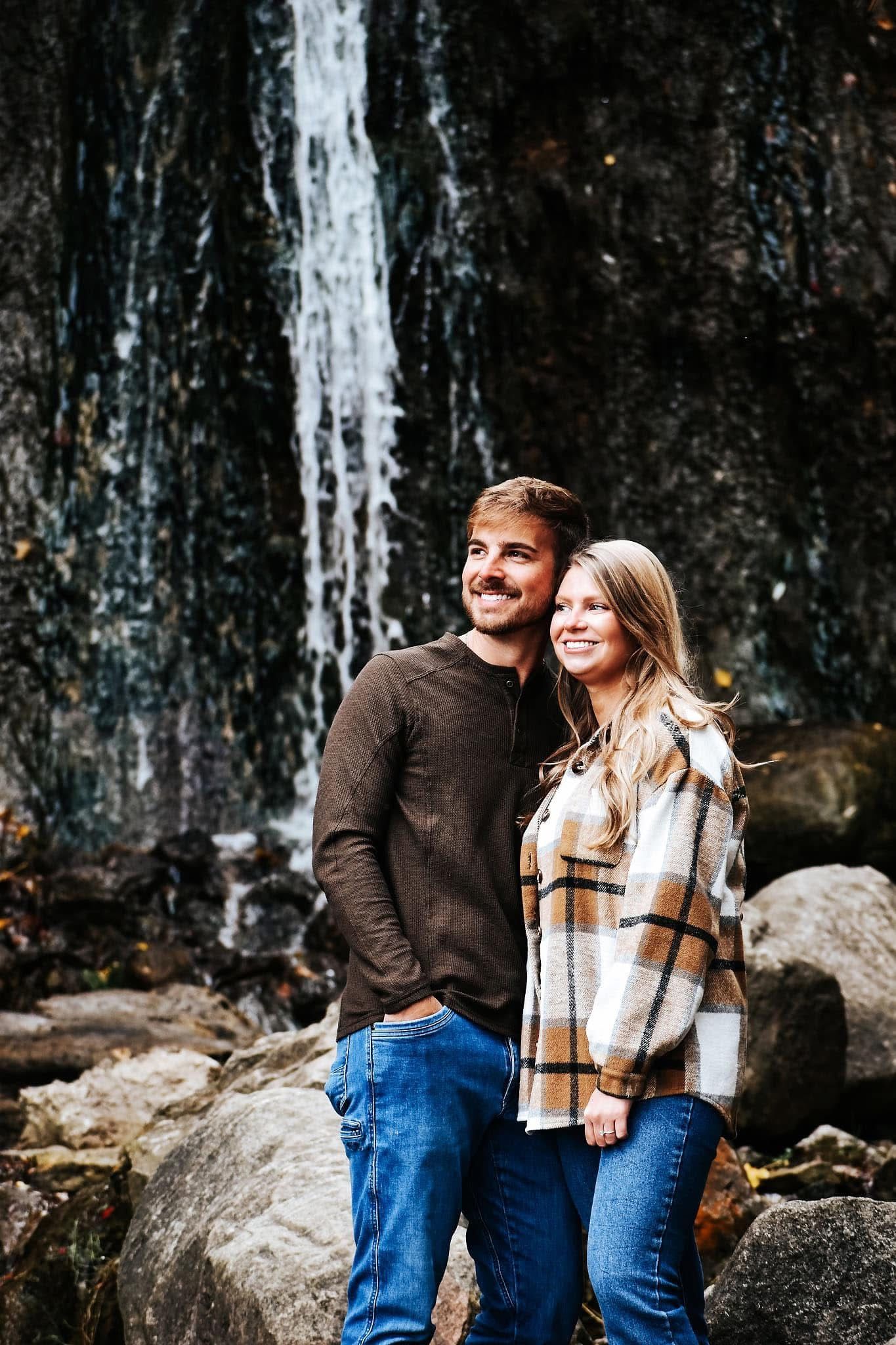 A man and a woman are standing in front of a waterfall.