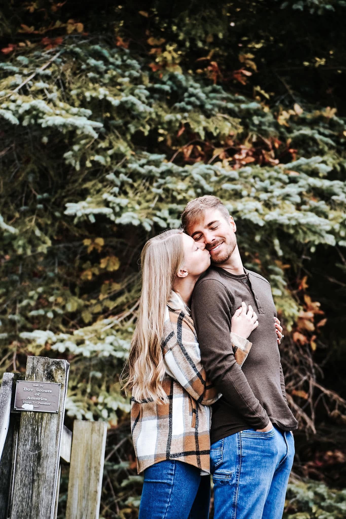 A woman is kissing a man on the cheek in front of trees.