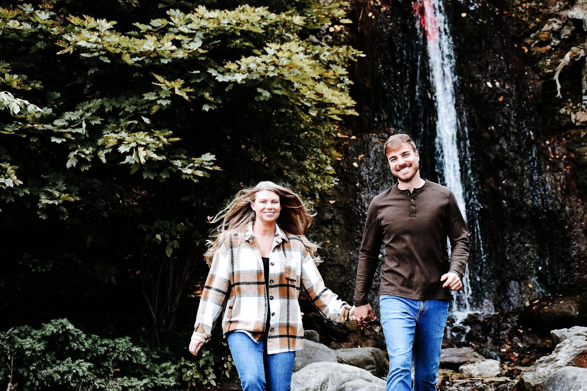 A man and a woman are walking next to a waterfall holding hands.