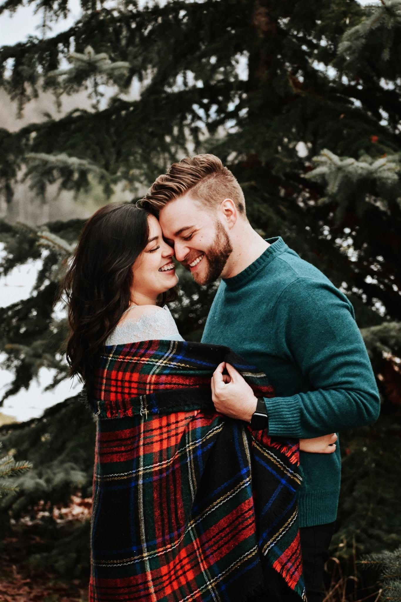 A man and a woman are standing next to each other wrapped in a plaid blanket.