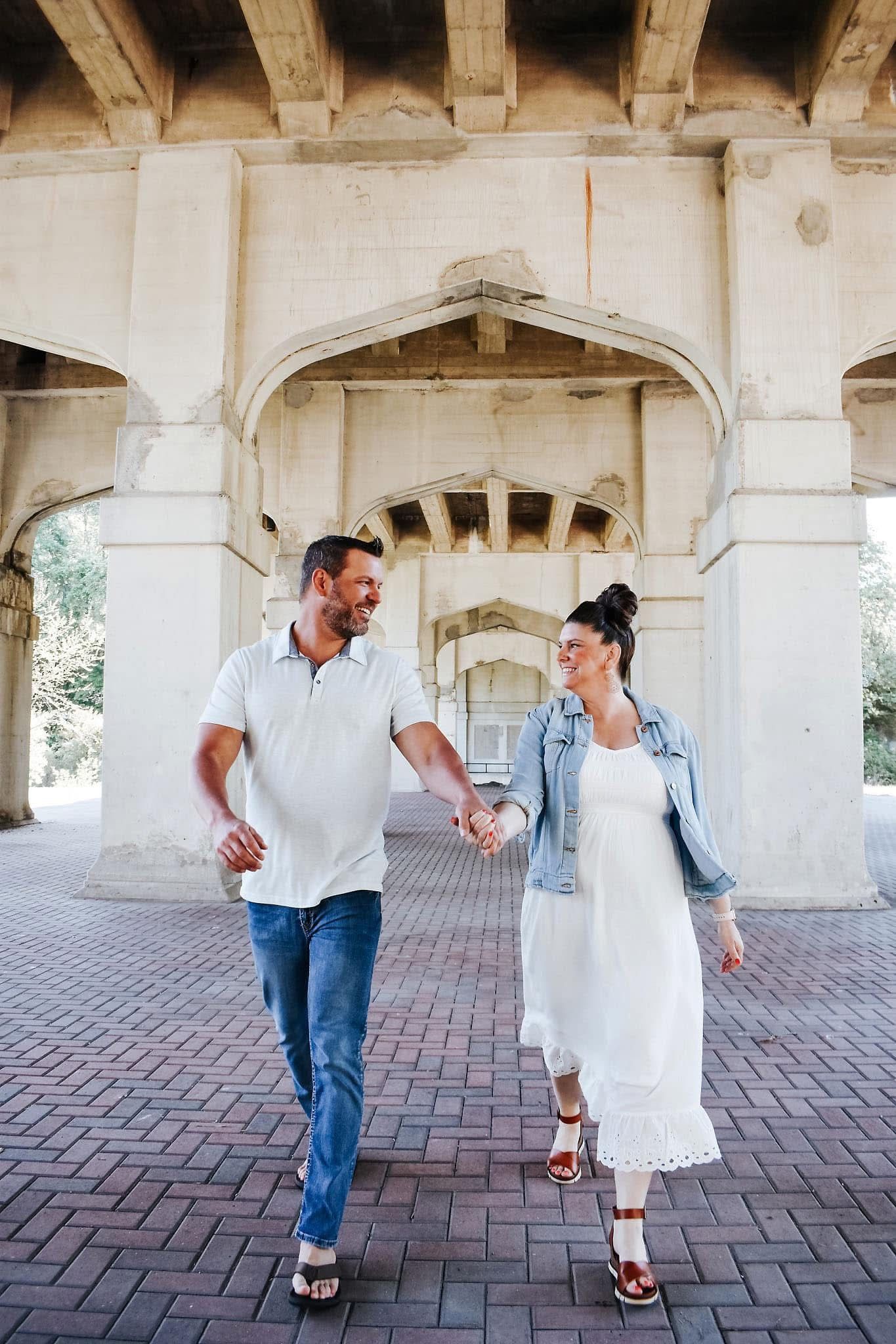 A man and a pregnant woman are walking under a bridge holding hands.