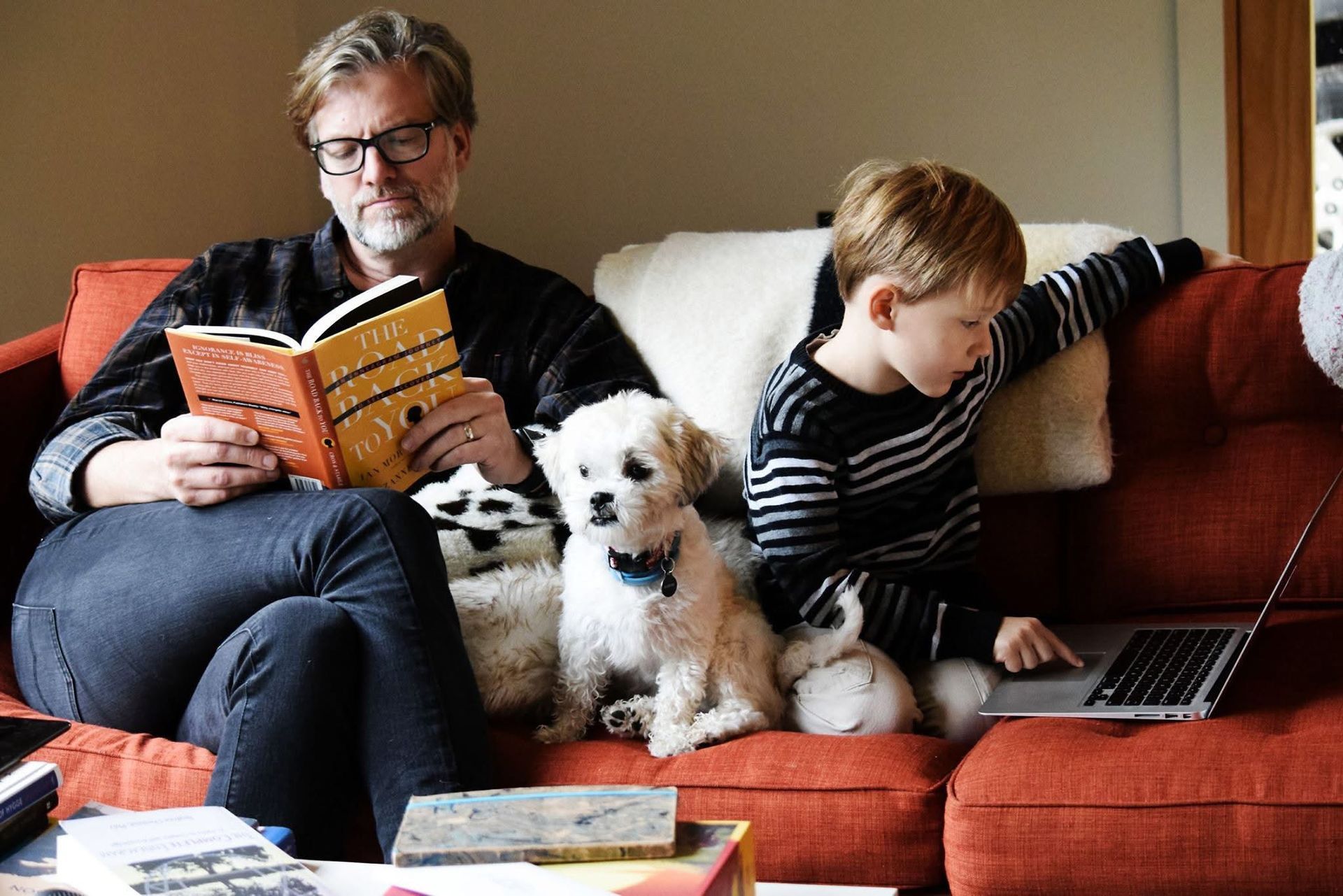A man and a boy are sitting on a couch with a dog and a laptop.