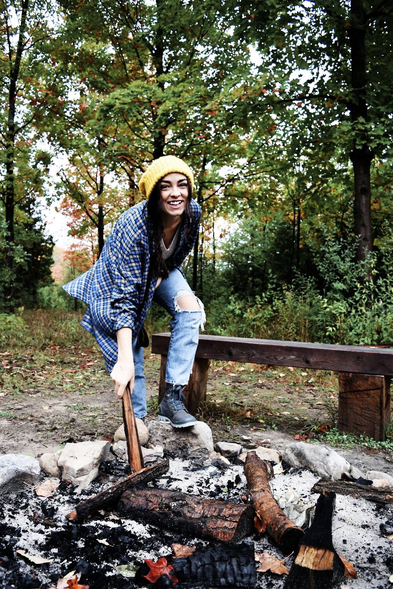 A woman is sitting on a bench next to a campfire in the woods.
