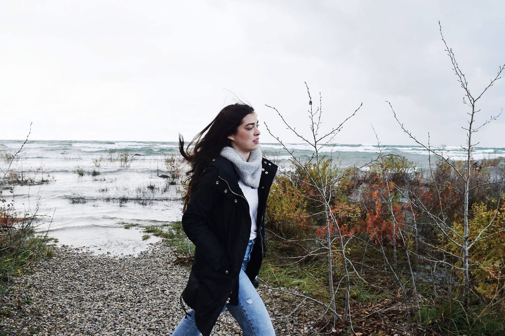 A woman in a black coat is walking on a path near the ocean.