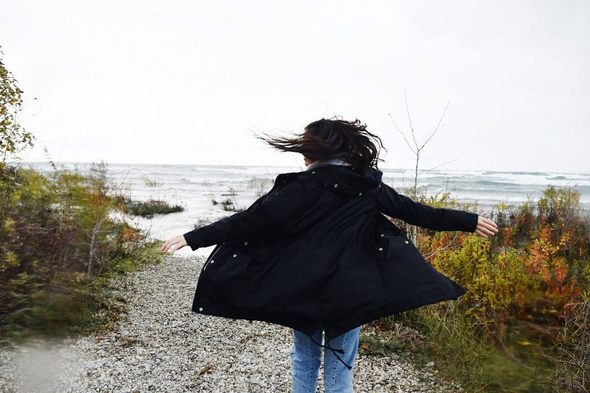 A woman in a black coat is running down a gravel path towards the ocean.