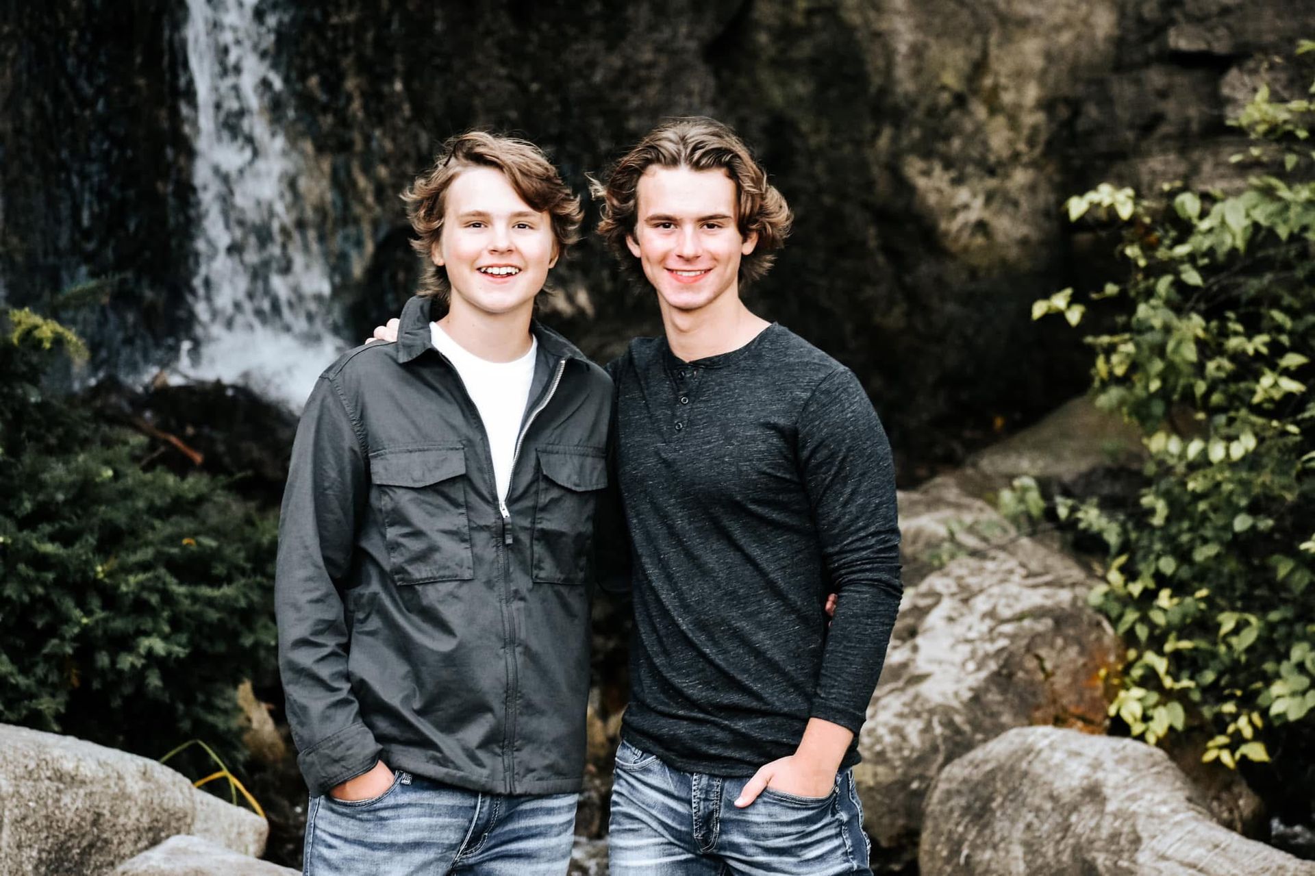 Two young men are standing next to each other in front of a waterfall.