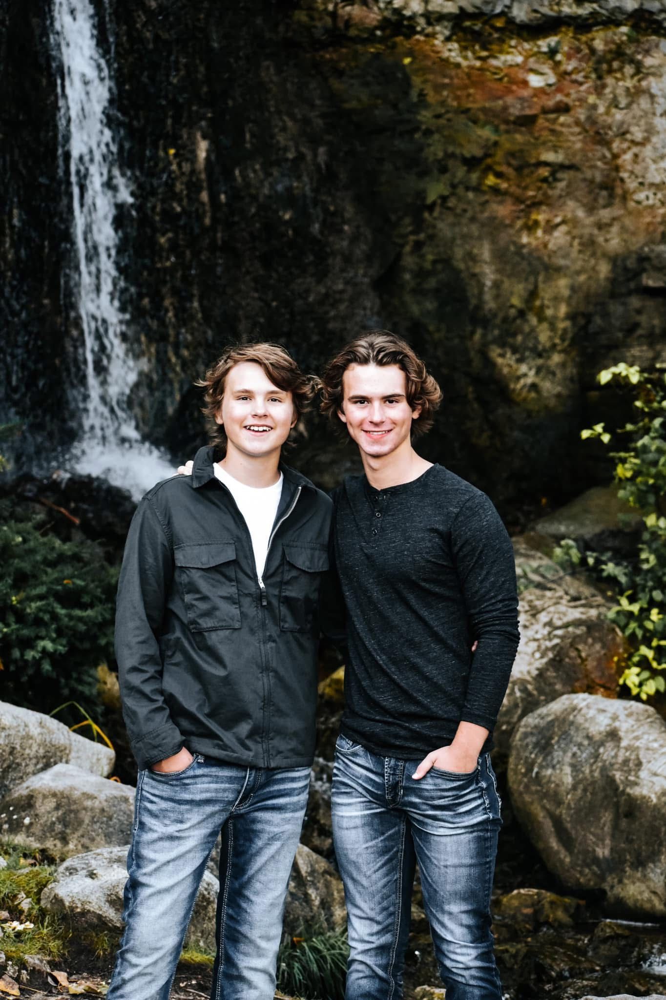 Two young men are posing for a picture in front of a waterfall.