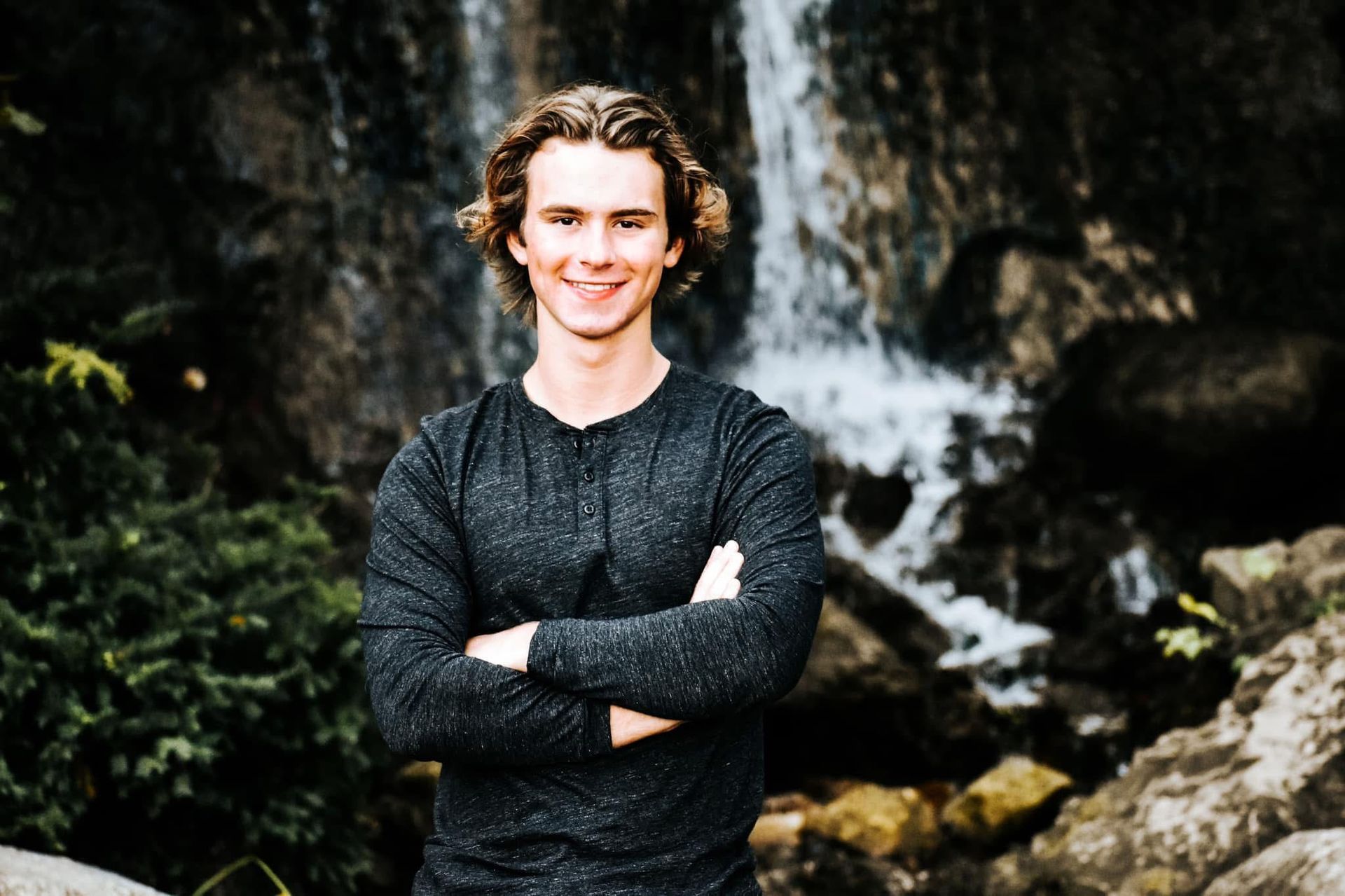 A young man is standing in front of a waterfall with his arms crossed.