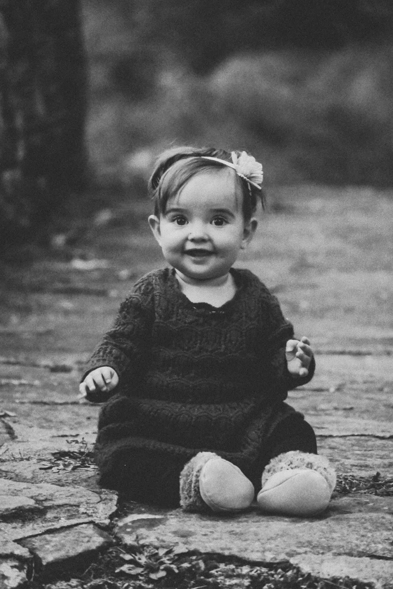 A black and white photo of a baby girl sitting on the ground.