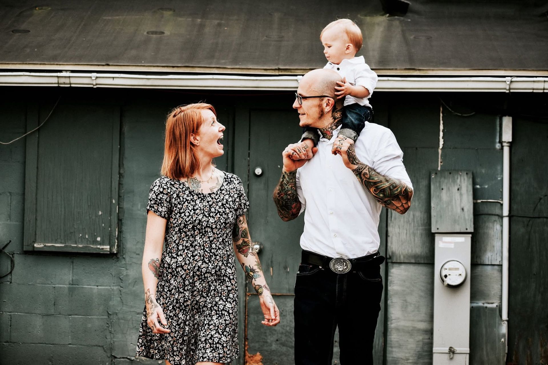 A man is carrying a baby on his shoulders while a woman looks on.