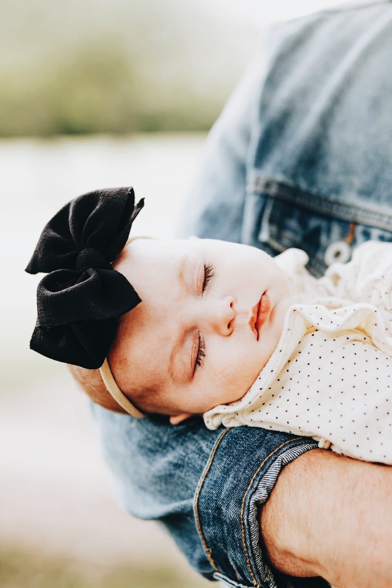 A baby with a black bow on her head is sleeping in a person 's arms.