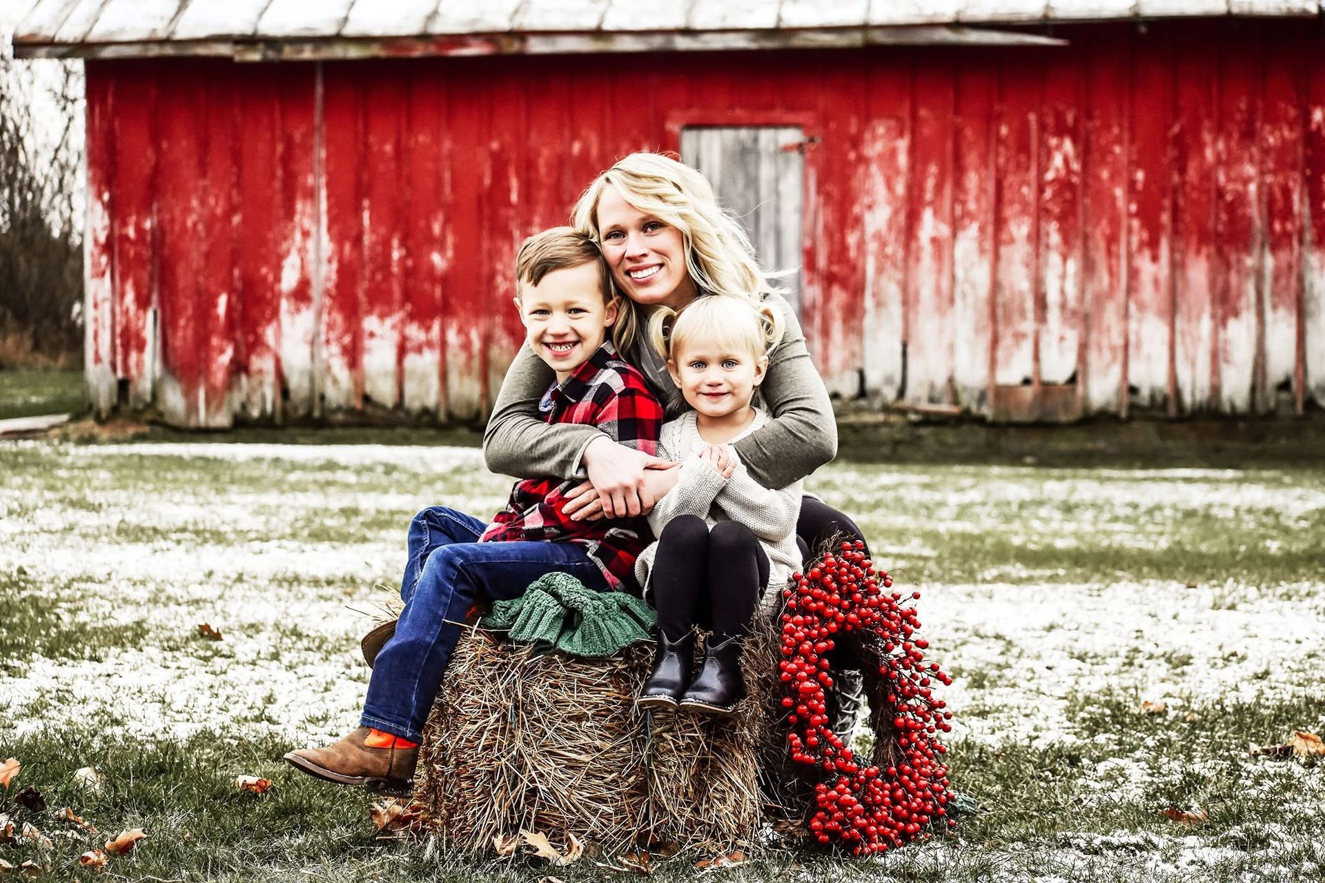 A woman and two children are sitting on a bale of hay in front of a red barn.