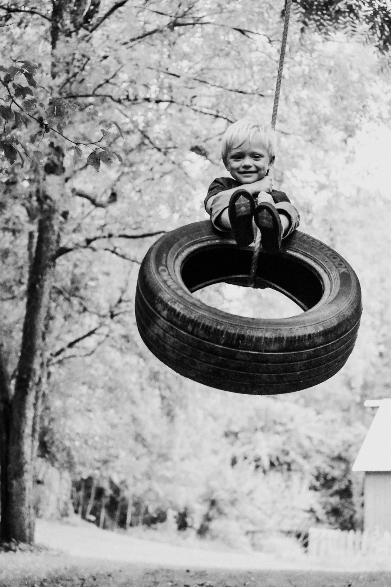 A black and white photo of a child sitting on a tire swing.