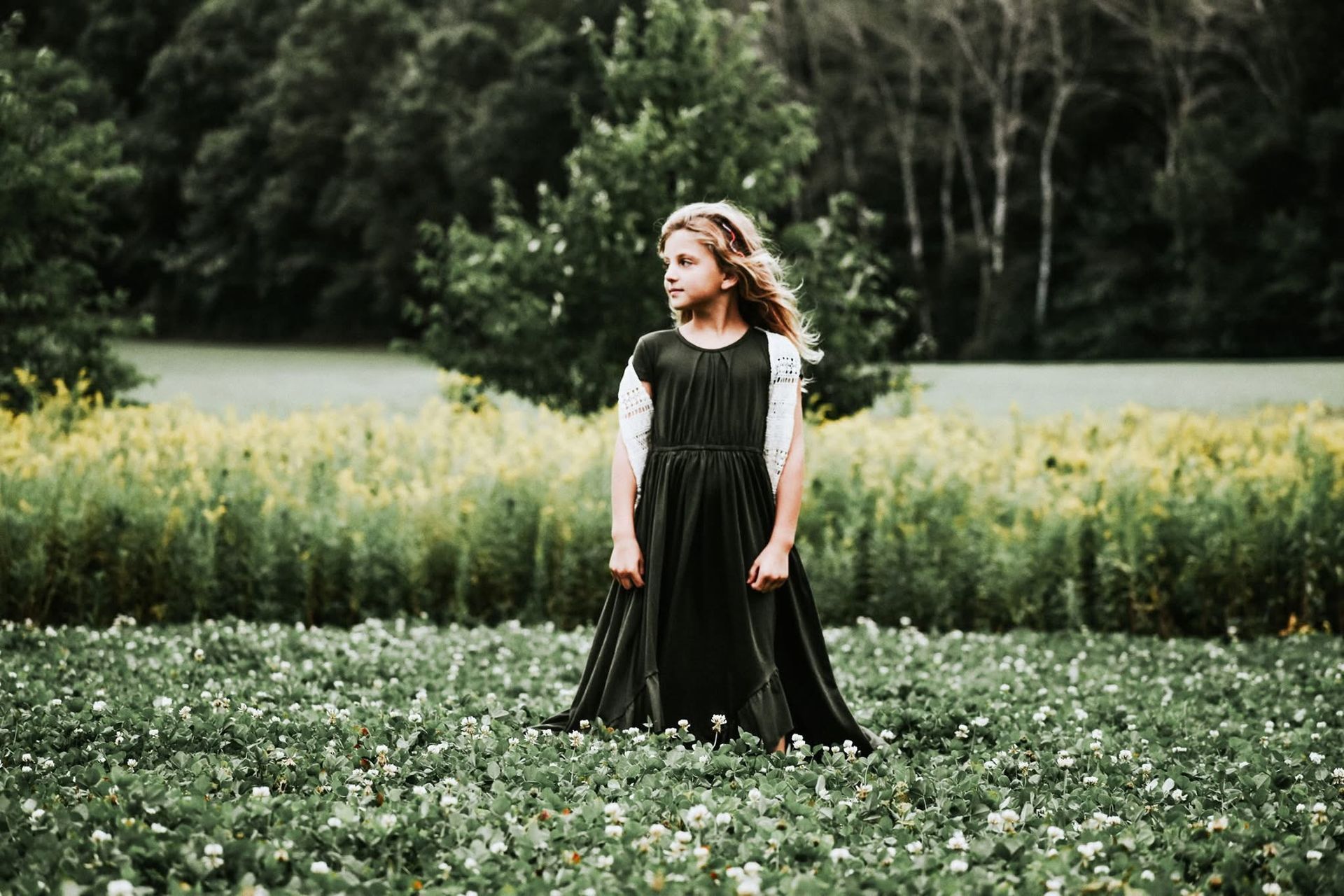 A little girl in a black dress is standing in a field of flowers.