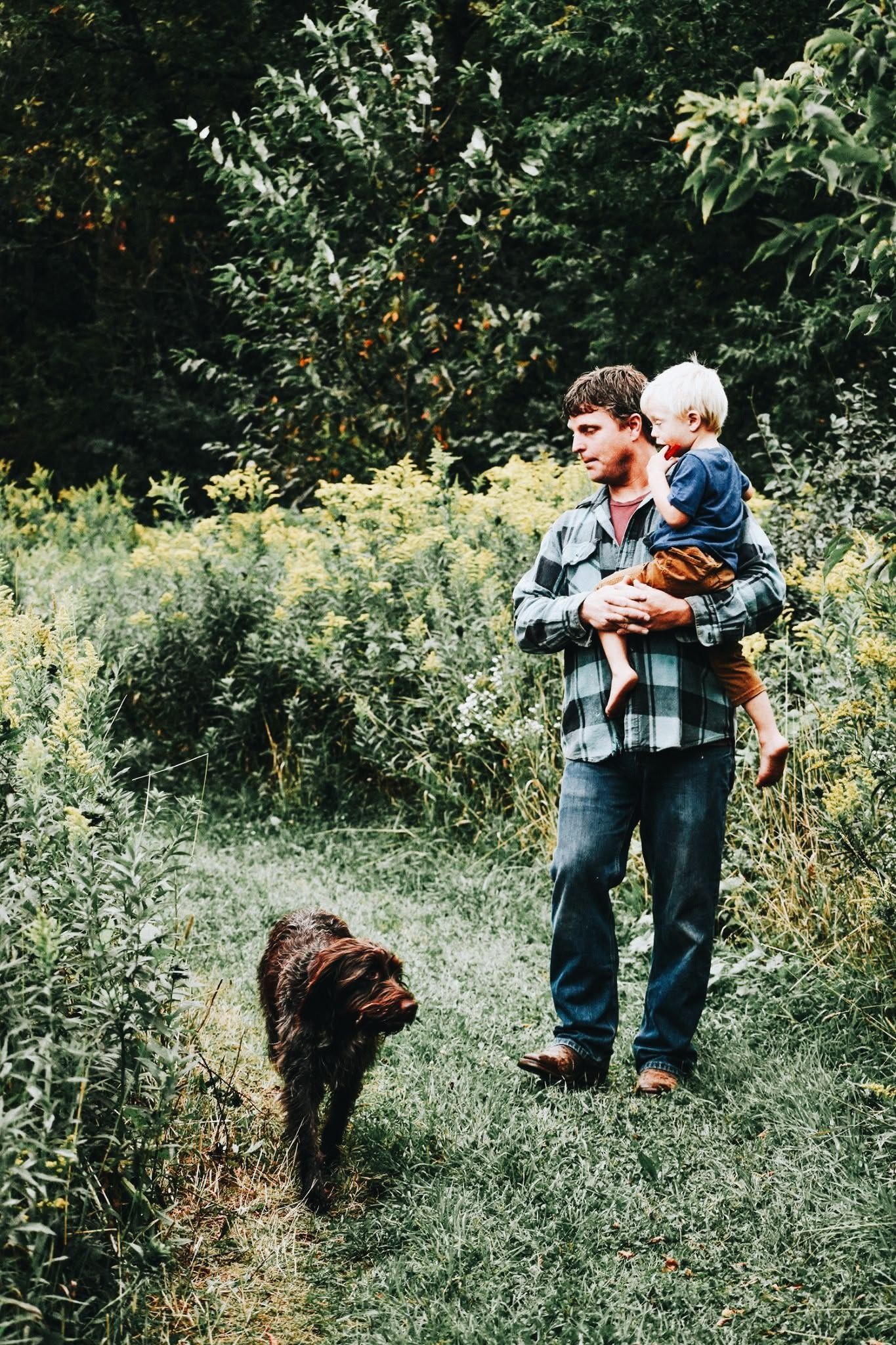 A man is holding a baby and a dog in a field