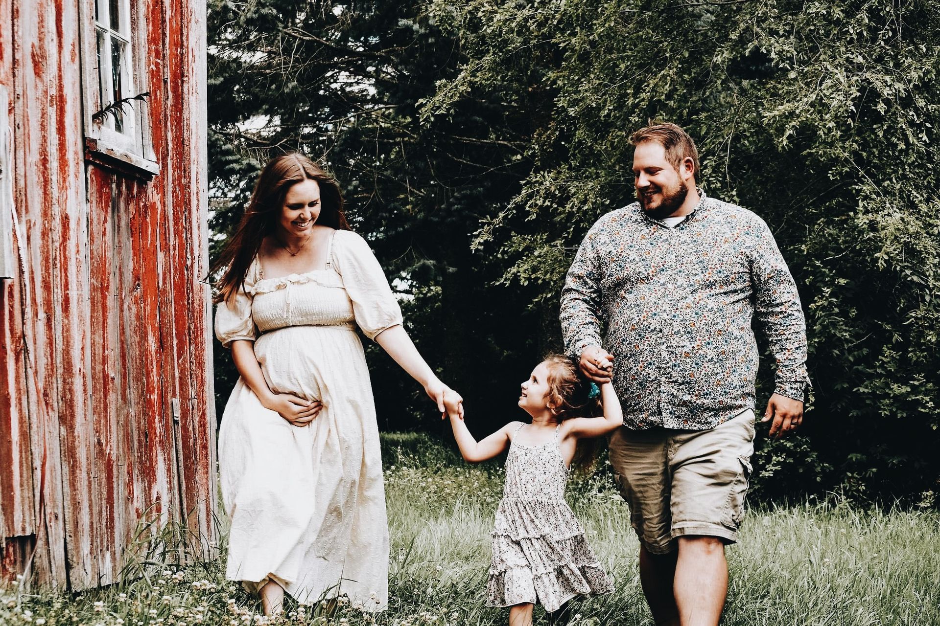 A pregnant woman , a man and a little girl are holding hands in front of a red barn.