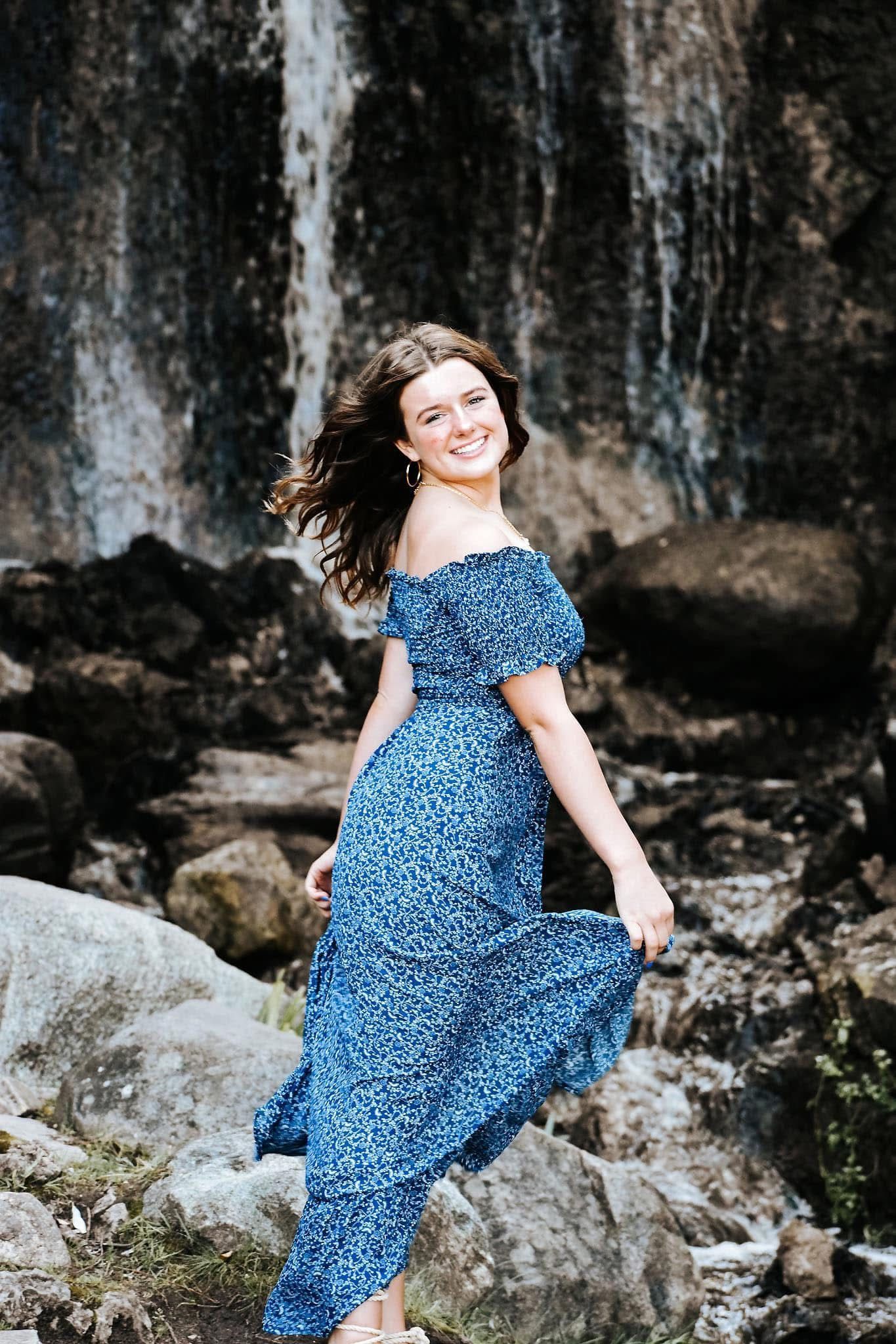 A woman in a blue dress is standing in front of a waterfall.