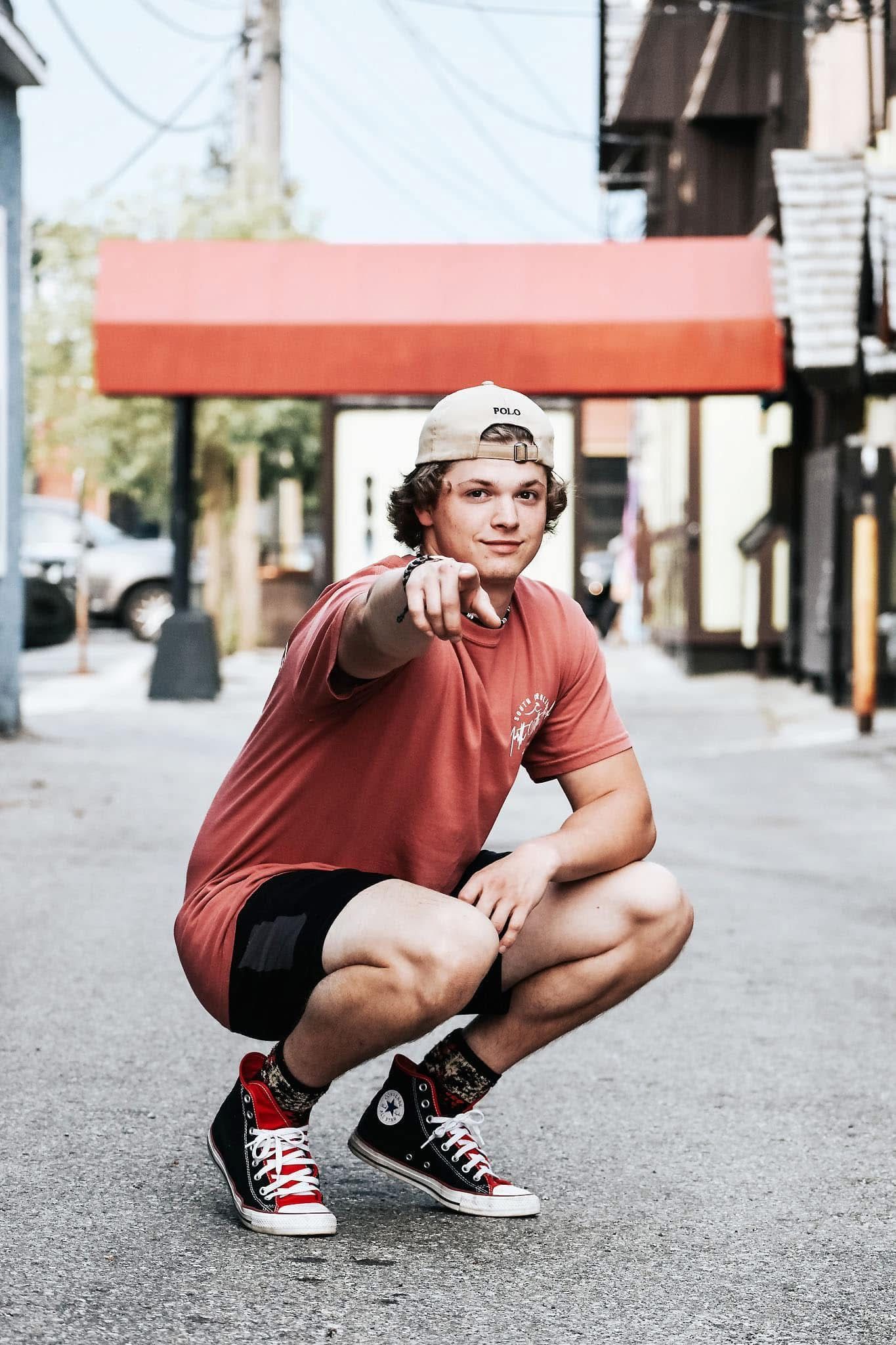 A young man is squatting down on the sidewalk and pointing at the camera.