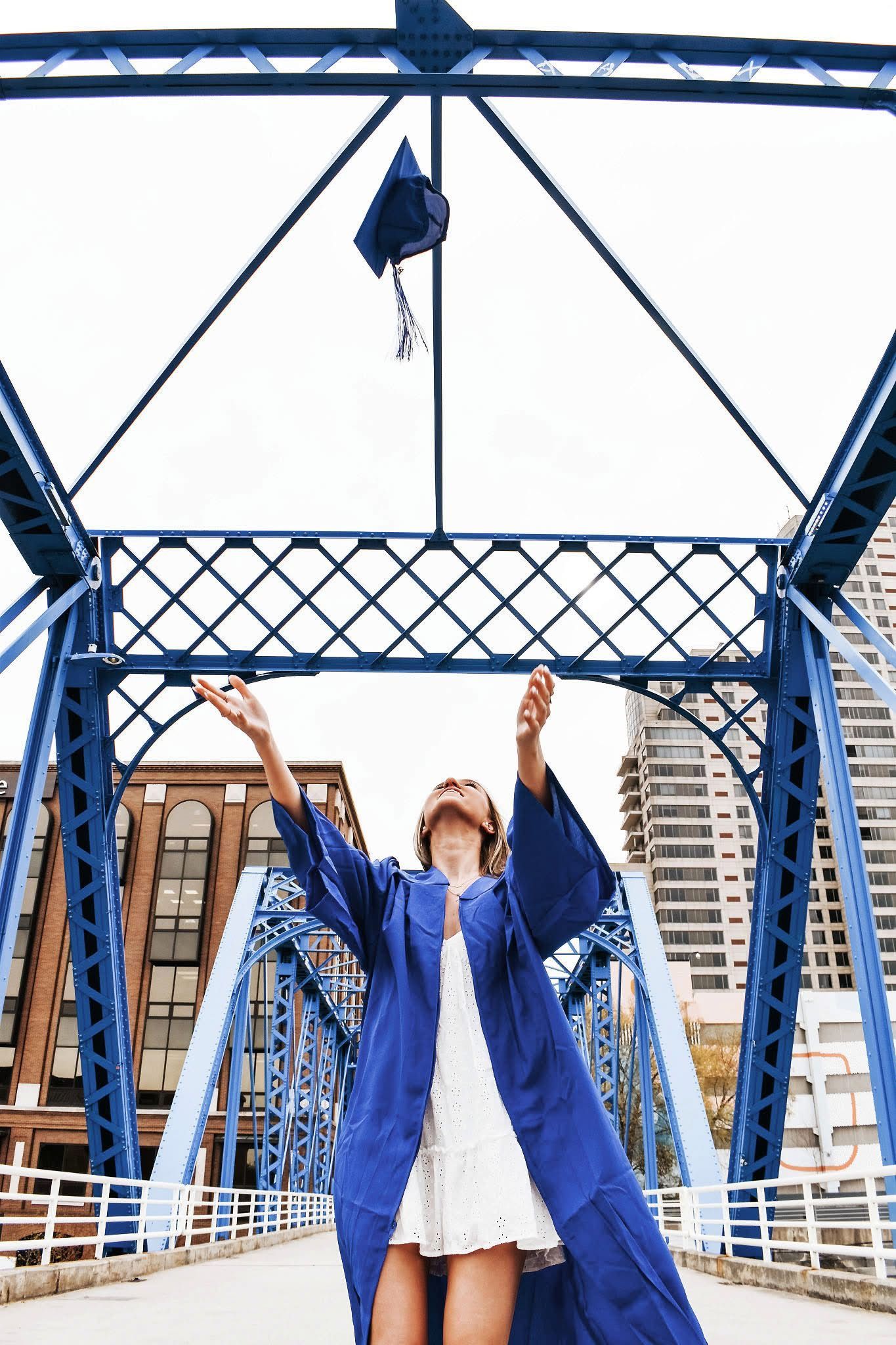 A woman in a blue cap and gown is throwing her graduation cap over a bridge.