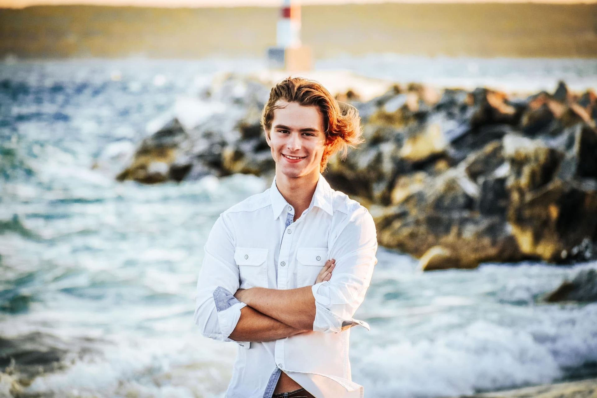 A young man in a white shirt is standing on the beach with his arms crossed.