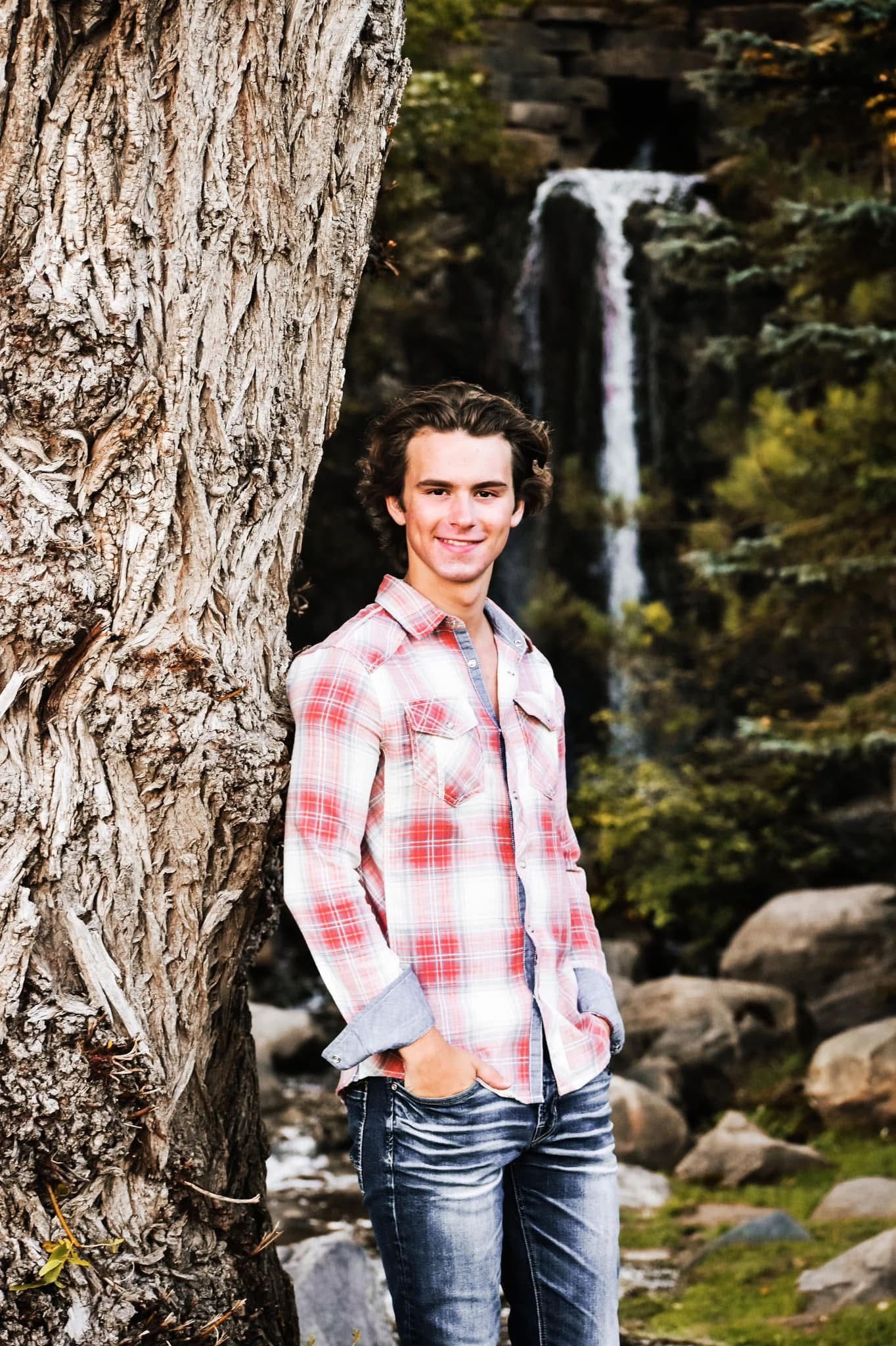 A young man is leaning against a tree in front of a waterfall.