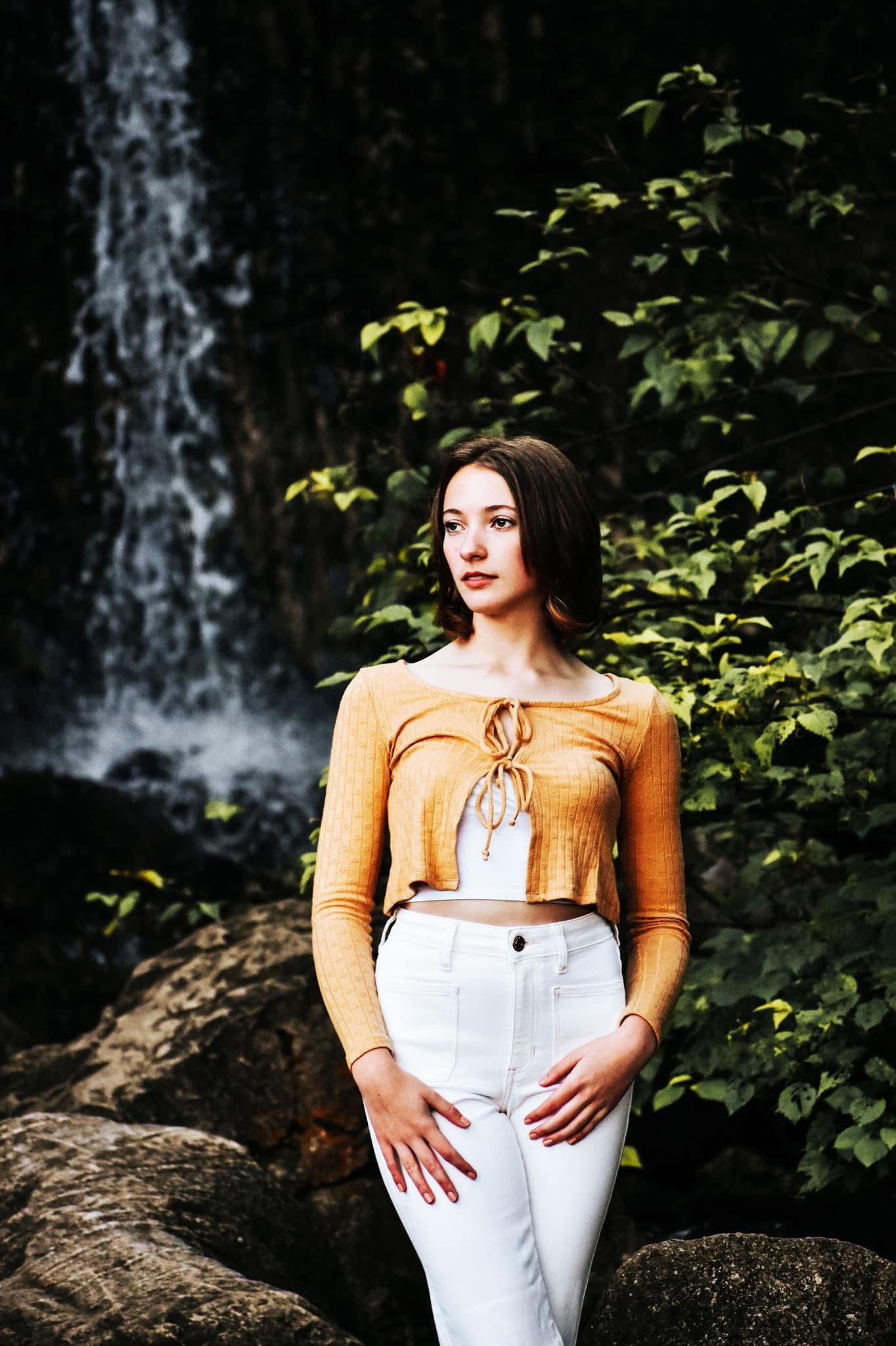 A woman in a yellow top and white pants is standing in front of a waterfall.