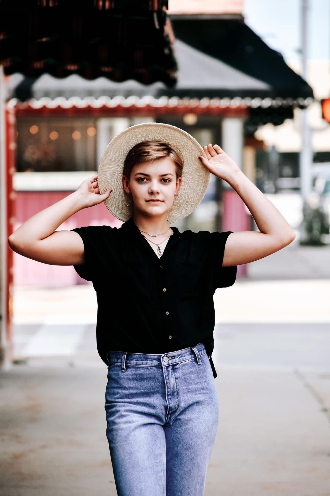 A woman wearing a straw hat and jeans is standing on a sidewalk.